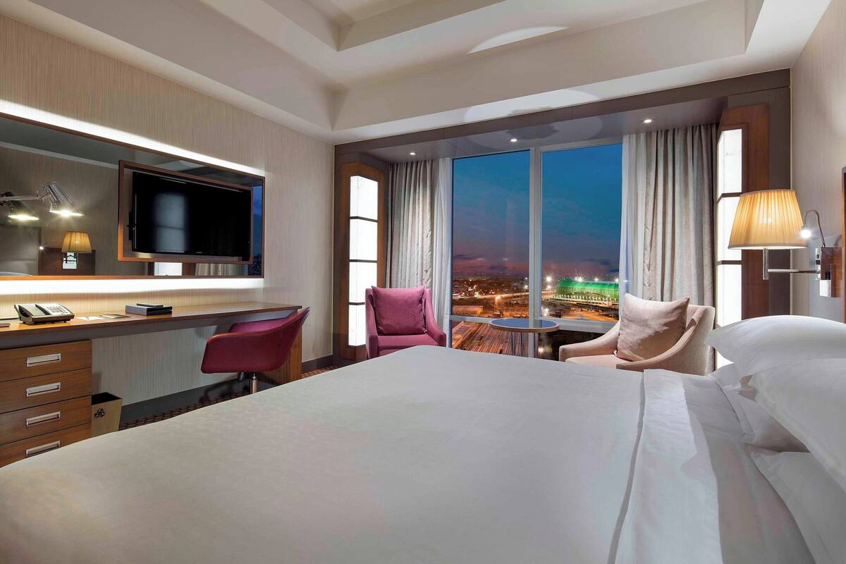 A hotel room with a large bed, a desk with a computer, a chair, a TV, and a view of the city at night.