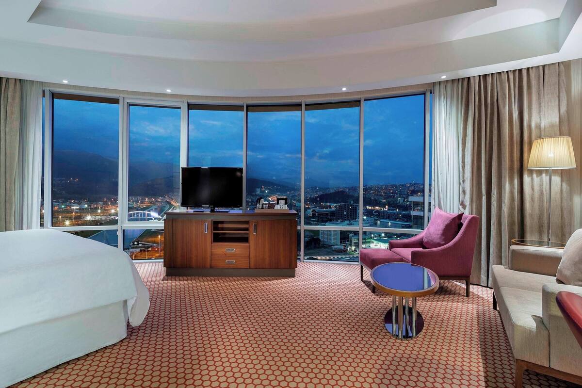 A hotel room with a large bed, a TV on a wooden cabinet, a pink chair, a round table, and a city view through large windows.
