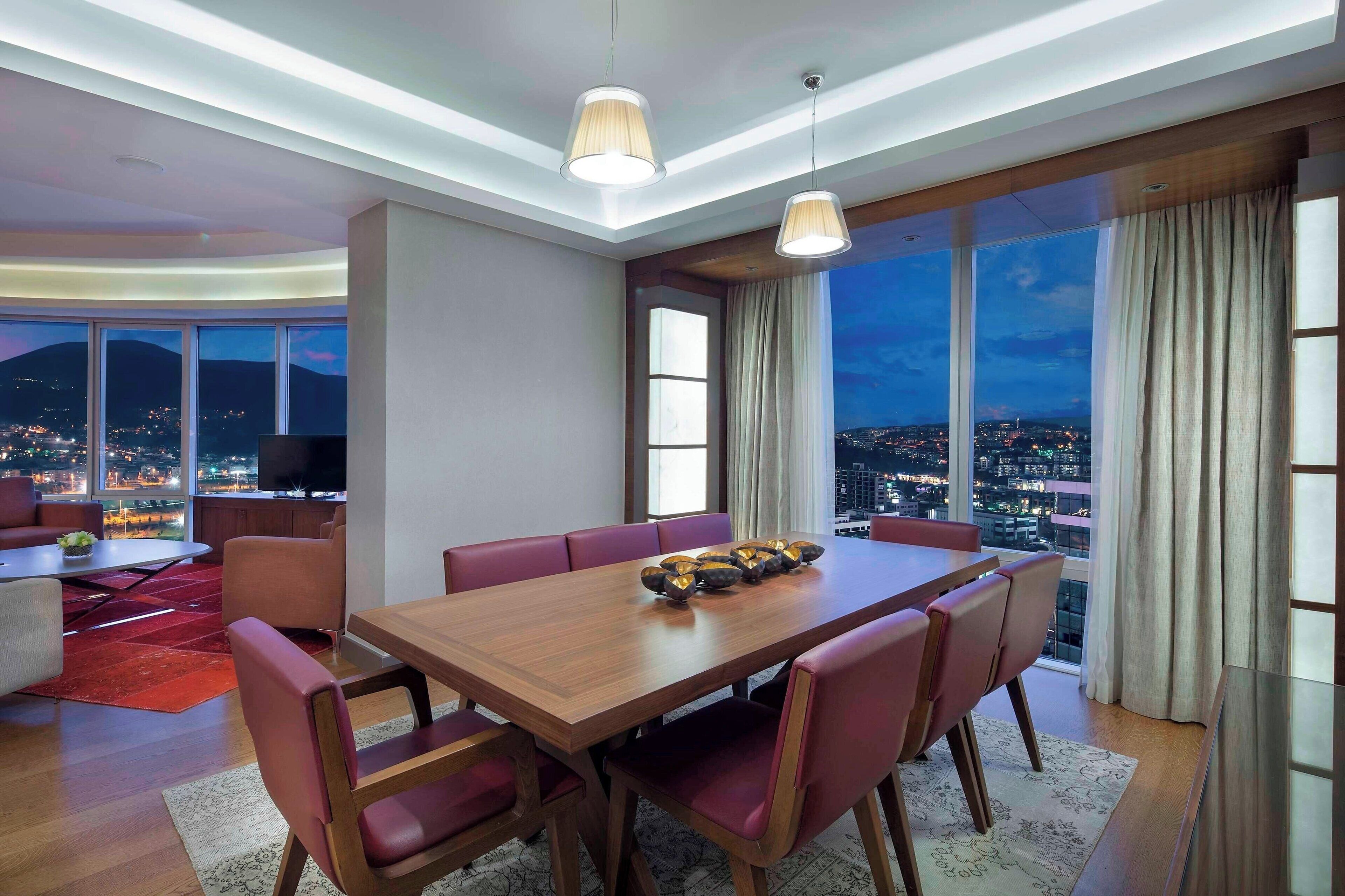A modern dining area with a wooden table and red upholstered chairs, large windows with a city view, and pendant lighting.