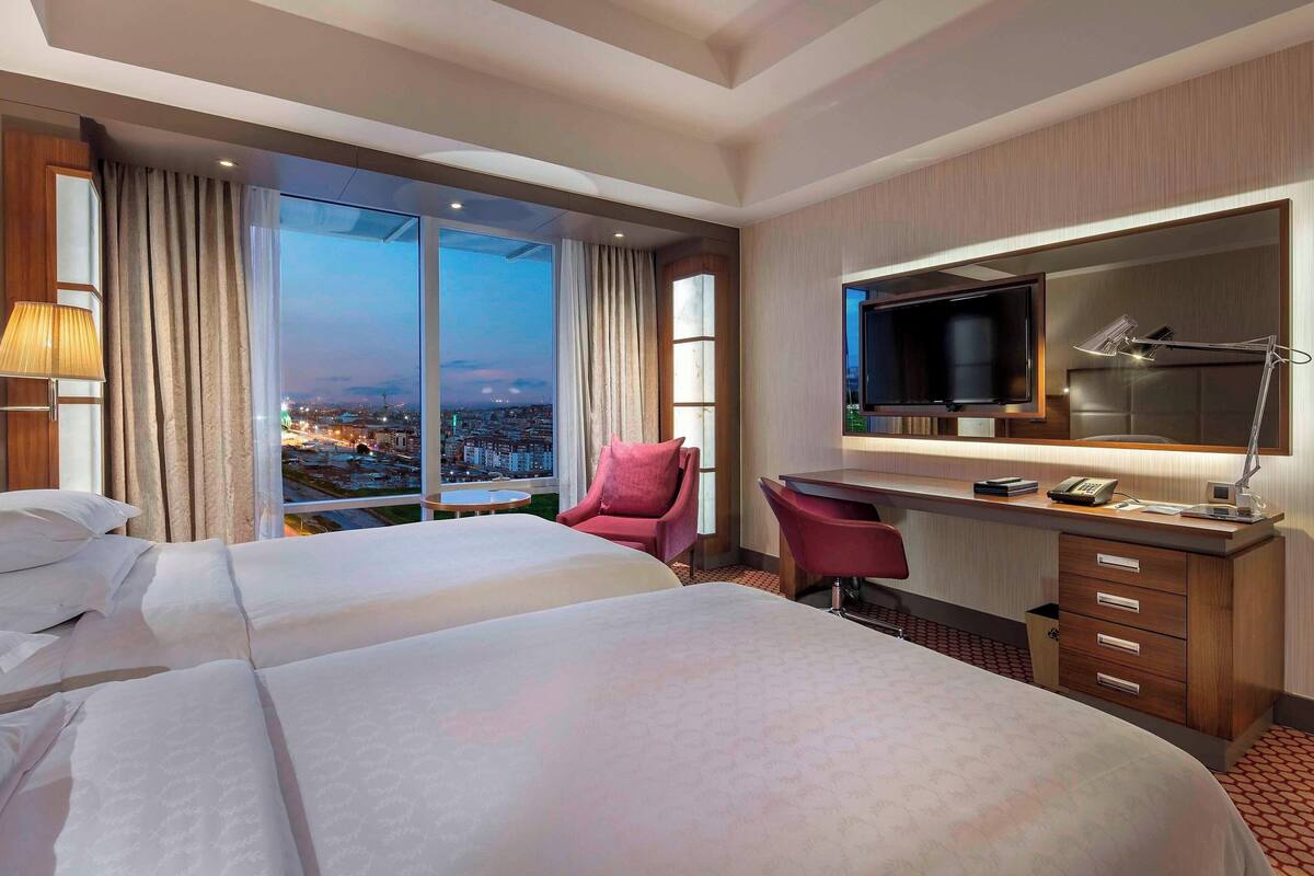 A hotel room with two beds, a desk with a TV, a chair, and a large window offering a city view.