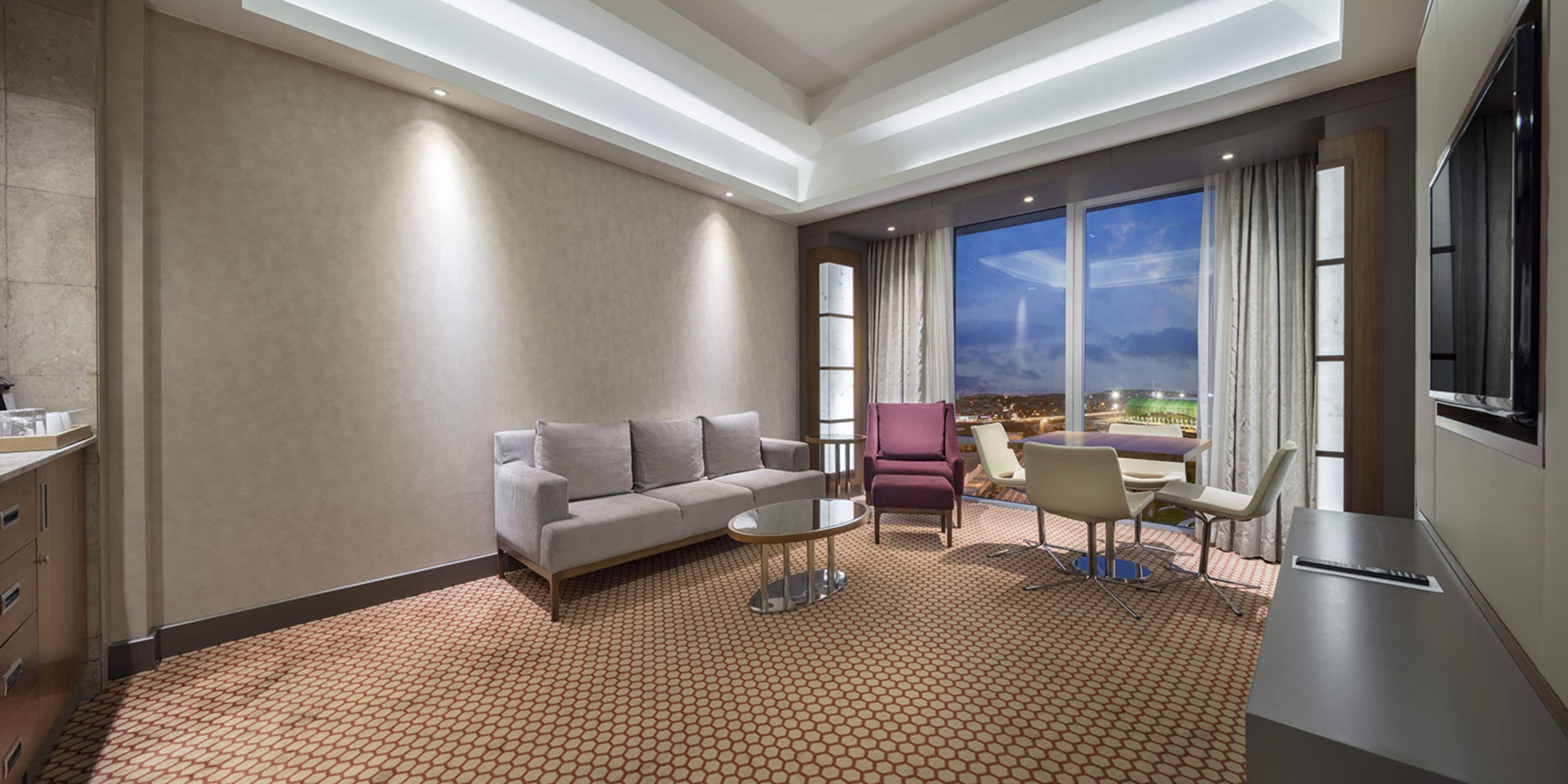 A modern hotel room with a sofa, armchairs, a small table, and a TV. The room has a large window with a view of the city.