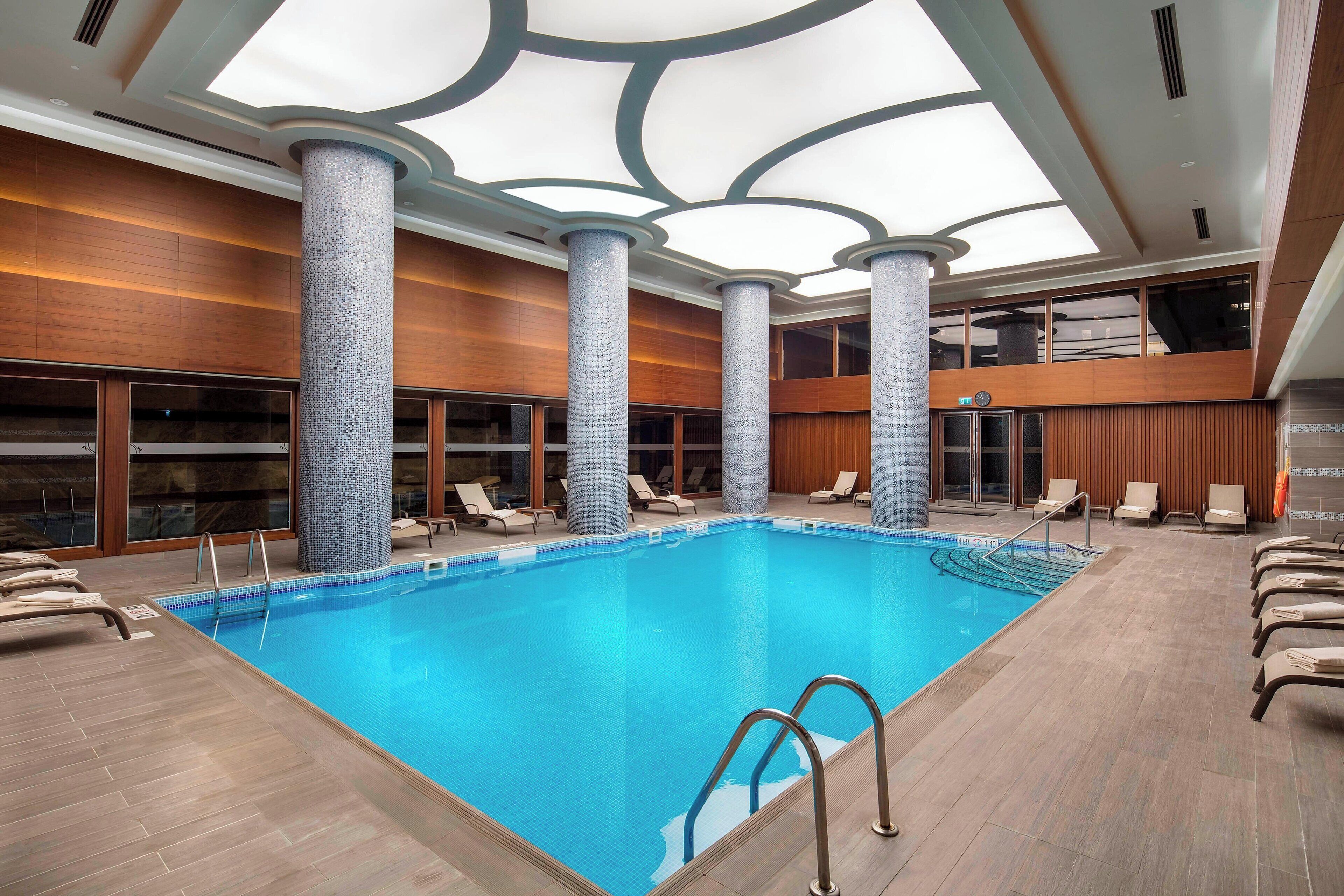Una piscina interior con agua azul cristalina, rodeada de sillones de descanso y un diseño de techo moderno.