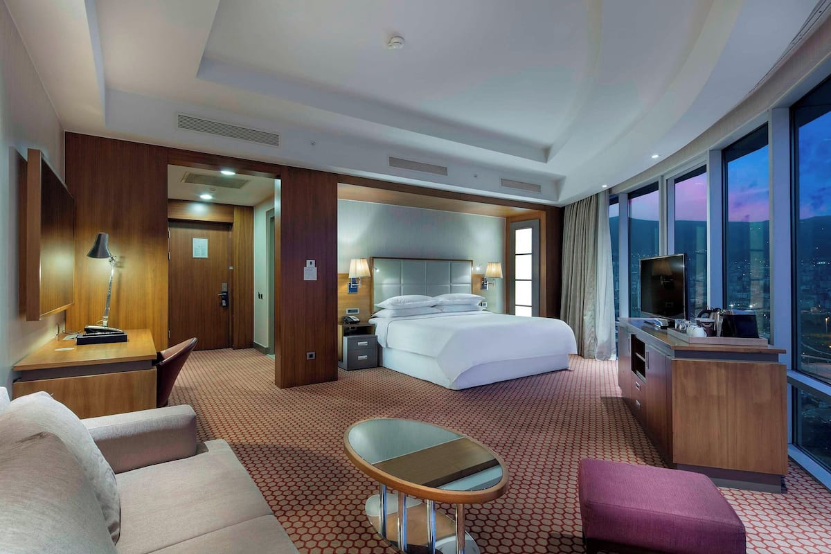 A hotel room with a bed, desk, and seating area, featuring a city view through large windows.