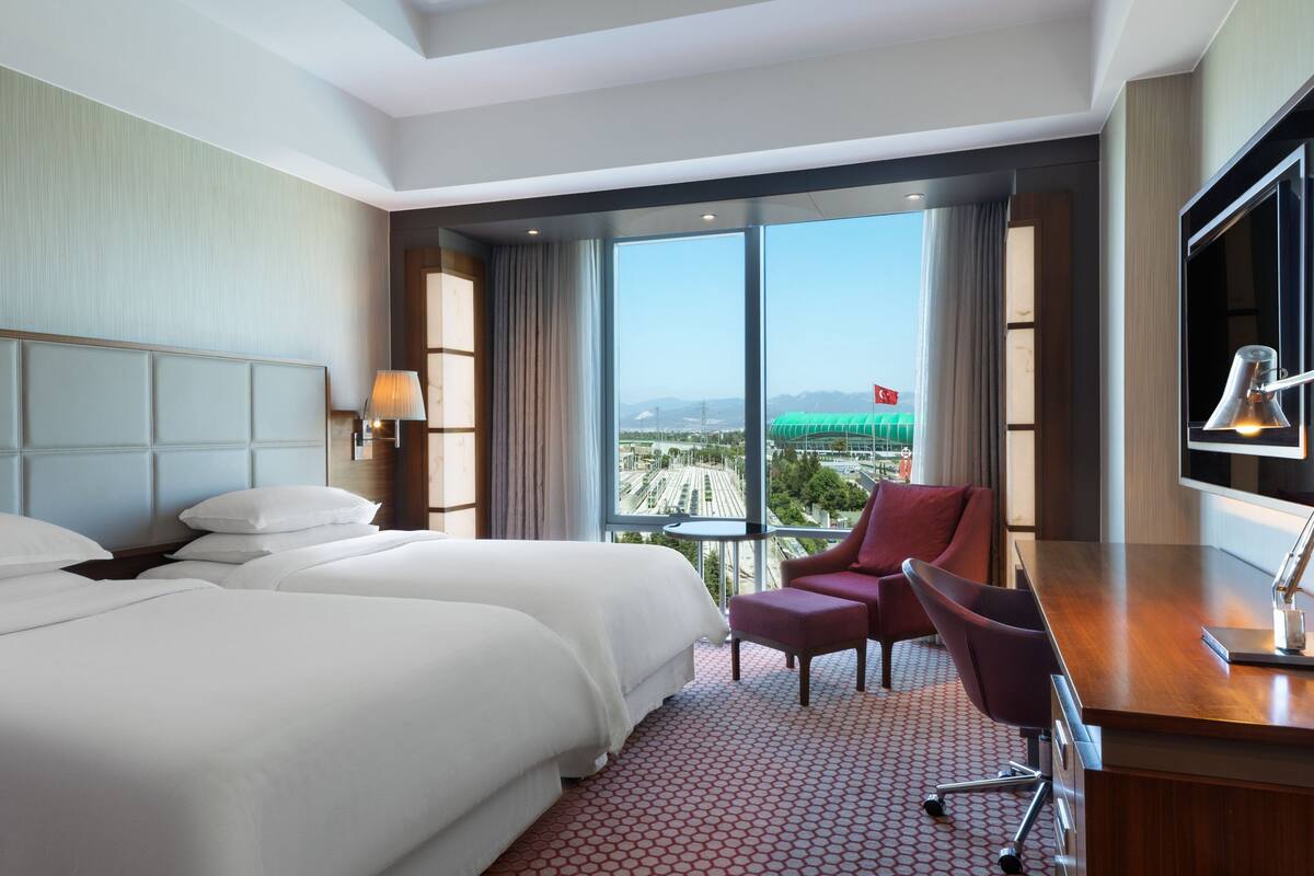 A hotel room with two beds, a desk, a chair, a TV, and a large window with a view of a cityscape and mountains.