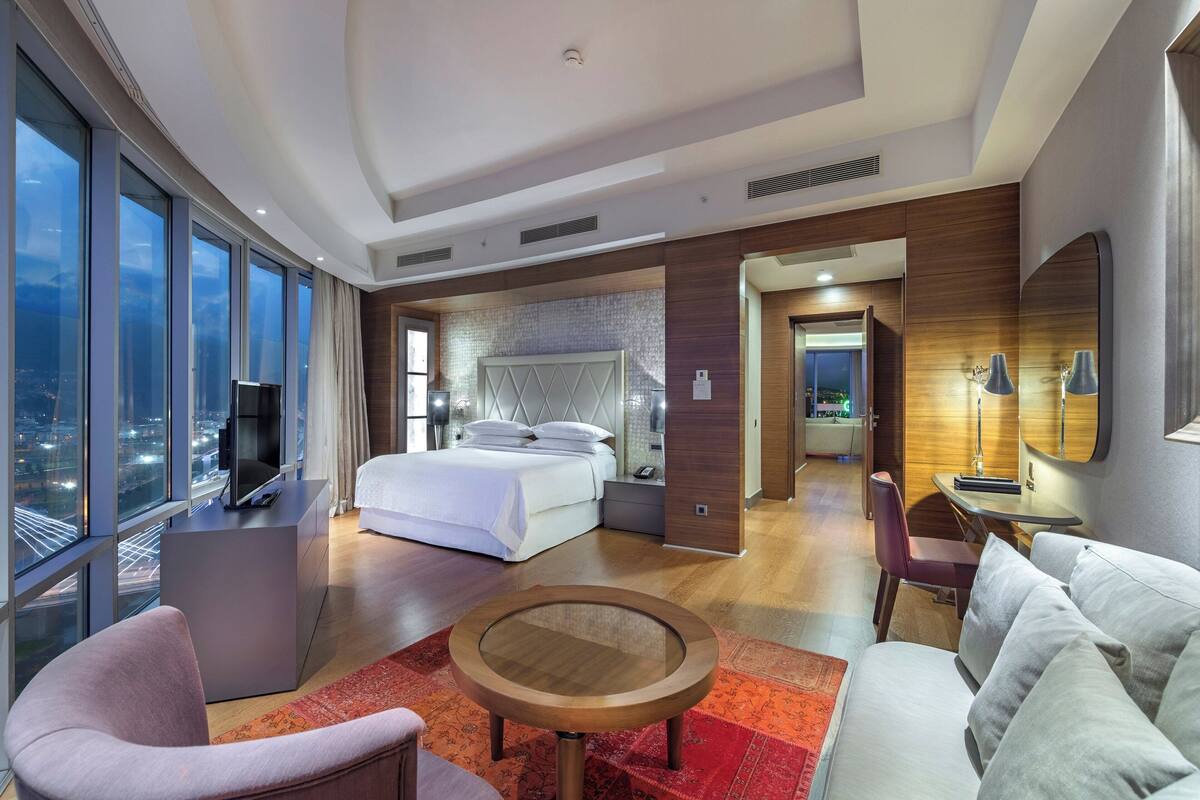 A modern hotel room with a large bed, a sitting area with a coffee table, a TV, and a city view through large windows.