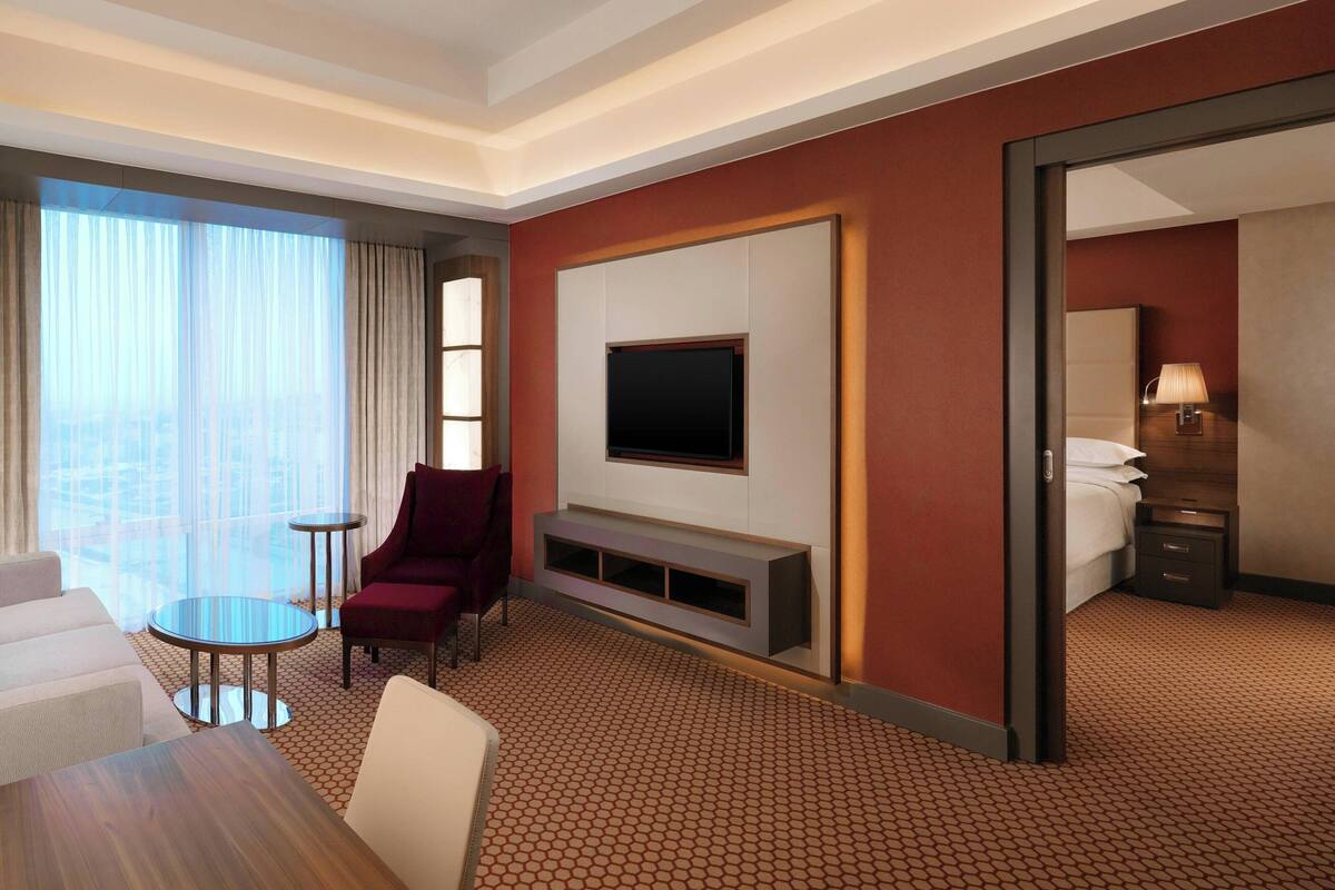 A hotel room with a large window, a flat-screen TV mounted on the wall, a seating area with a table and chairs, and a bed with a nightstand.