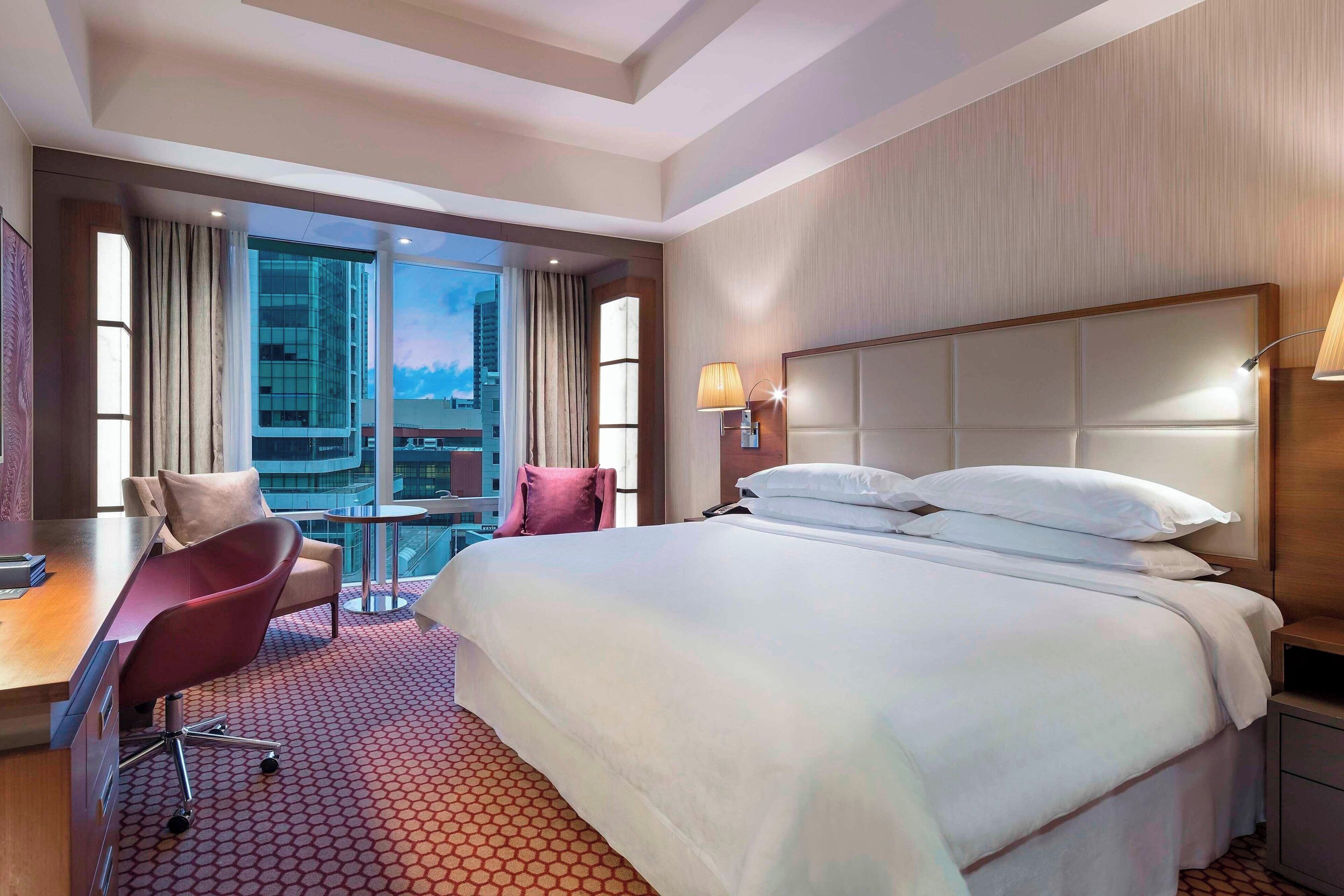A hotel room with a large bed, a desk with a chair, a small table, and a view of a cityscape through large windows.