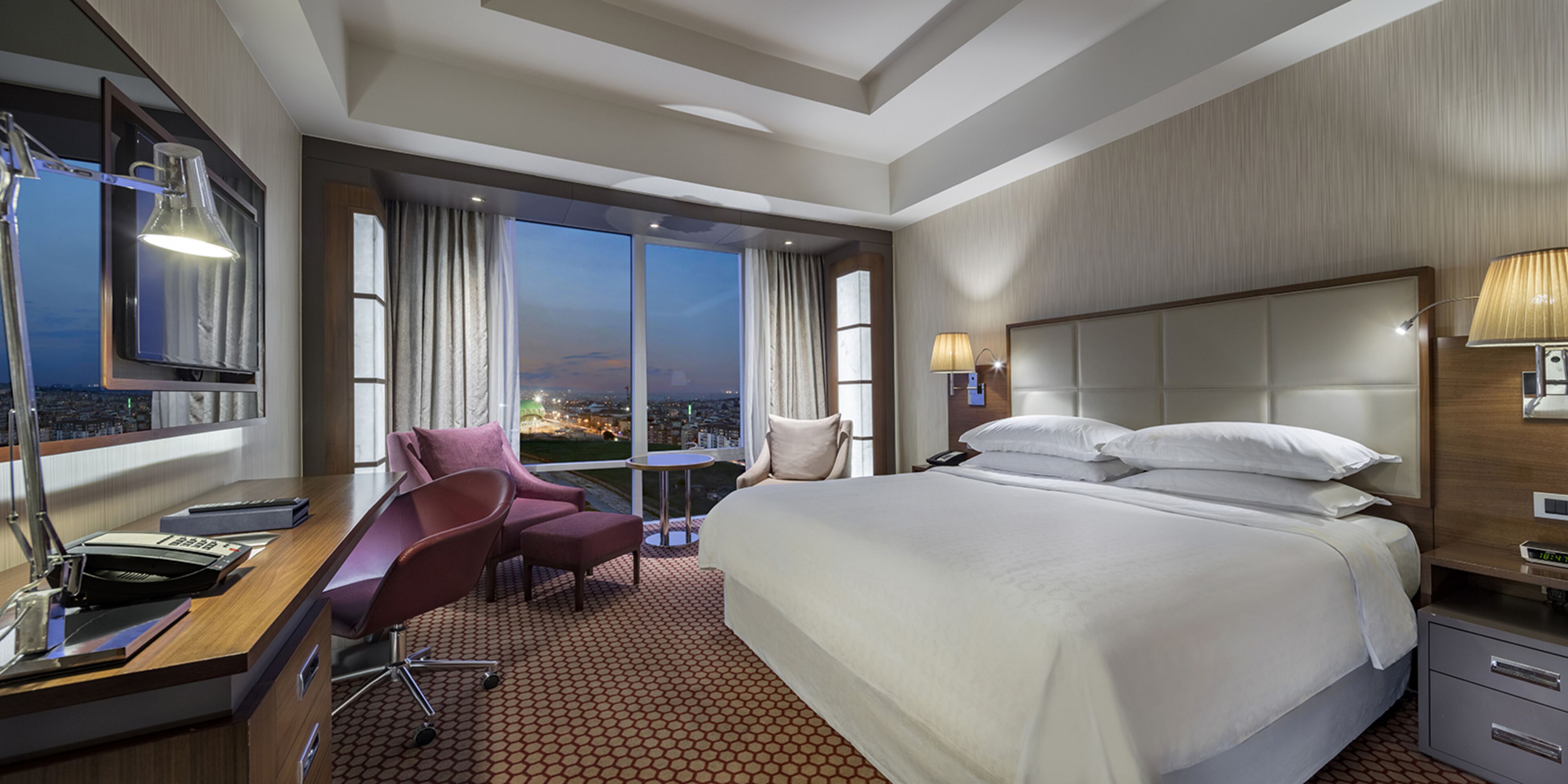 A hotel room with a large bed, a desk with a chair, a small table, and a view of the city at dusk.