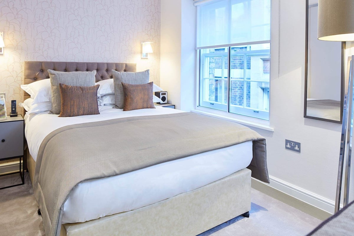 A neatly arranged bedroom with a large bed, two bedside tables, a nightstand, a window with a view, and a wall-mounted lamp.