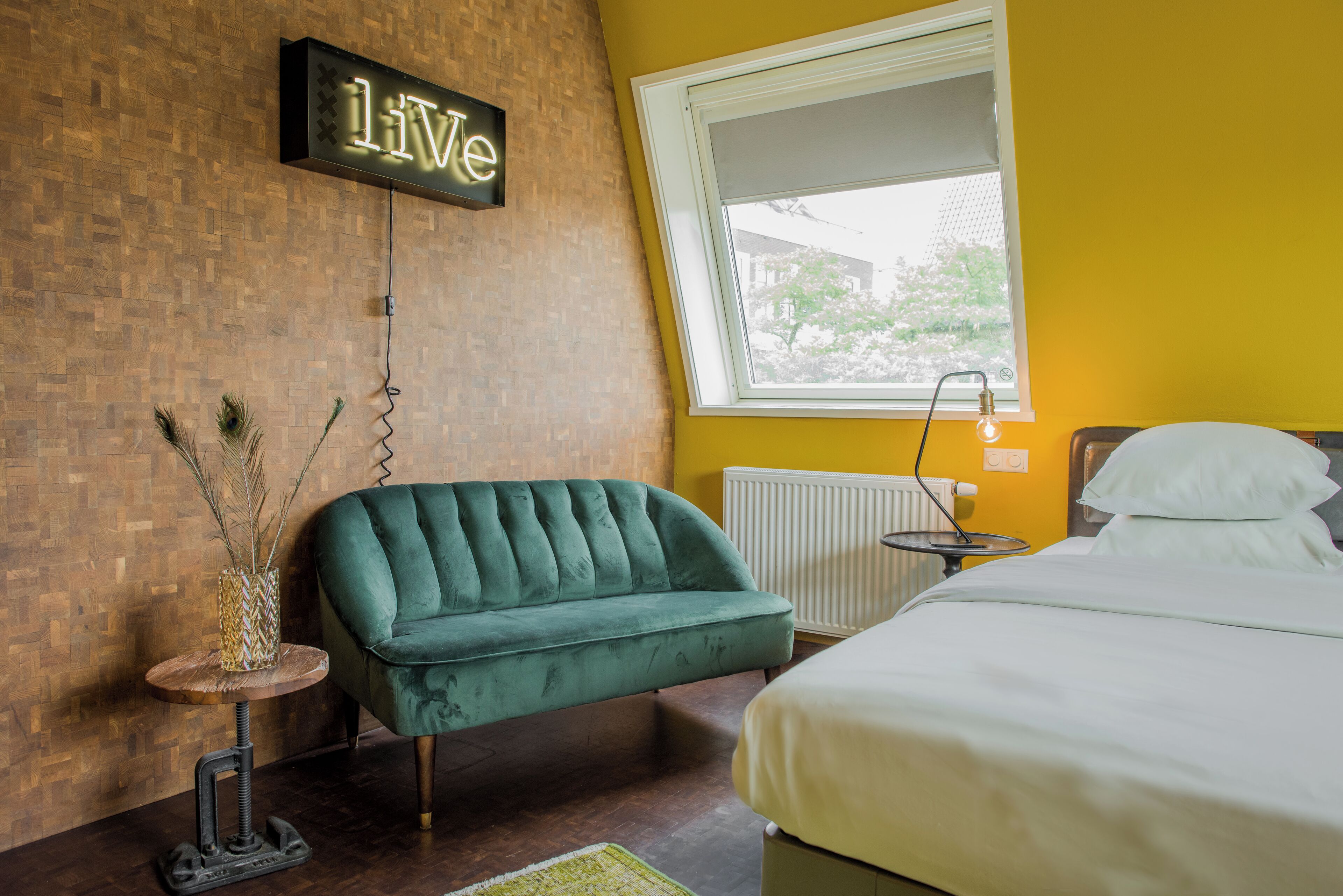 Una habitación con un sofá verde, una cama con ropa de cama blanca, una mesita con una lámpara y un cartel que dice 'Live' en la pared.