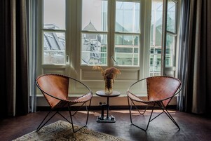 A room with two leather chairs, a small table with a vase, and a view of buildings through large windows.