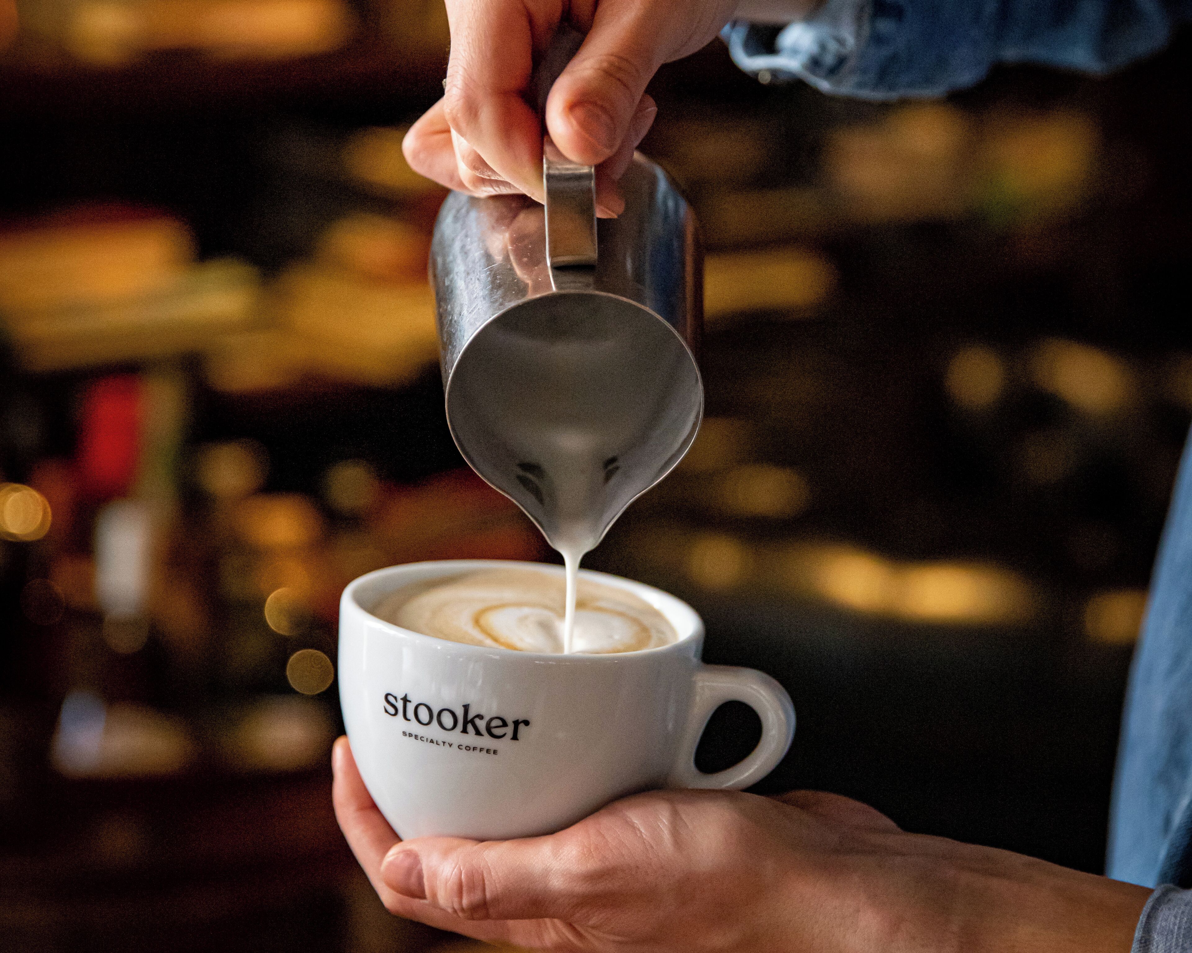 En person häller mjölk i en kopp som är märkt med texten ”stooker – specialkaffe”.