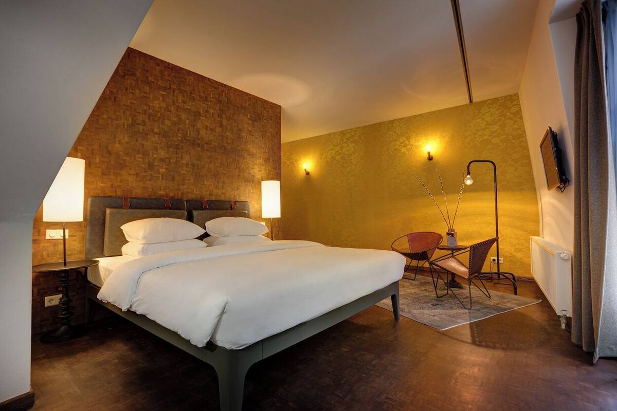 A modern hotel room with a large bed, a desk, and a chair. There are wall sconces providing light.