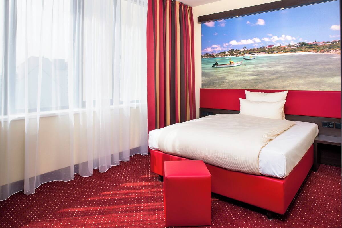 A hotel room with a large bed, a red ottoman, a view of a beach with boats, and sheer curtains.