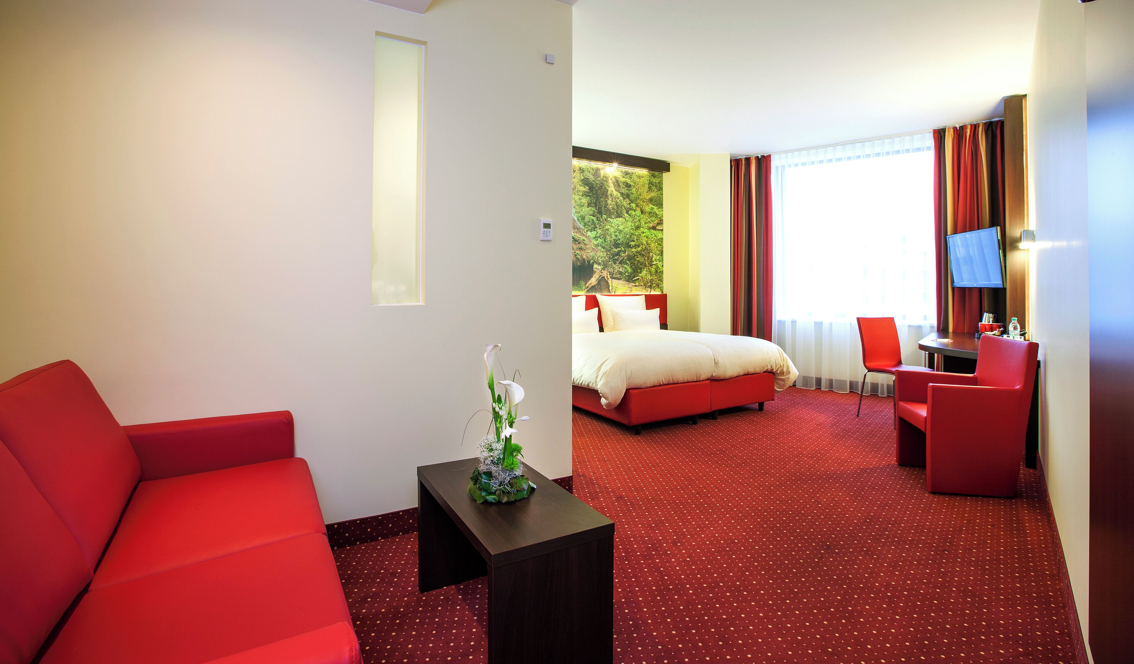 Een hotelkamer met een rode bank, een klein tafeltje met een plant, een bed met rood beddengoed, een bureau met een stoel en een televisie.