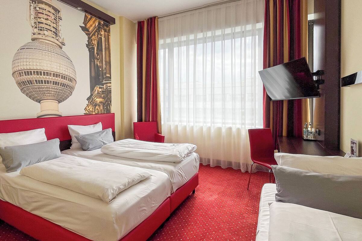 A hotel room with two beds, a red headboard, a large window with sheer curtains, a red chair, and a wall-mounted TV.