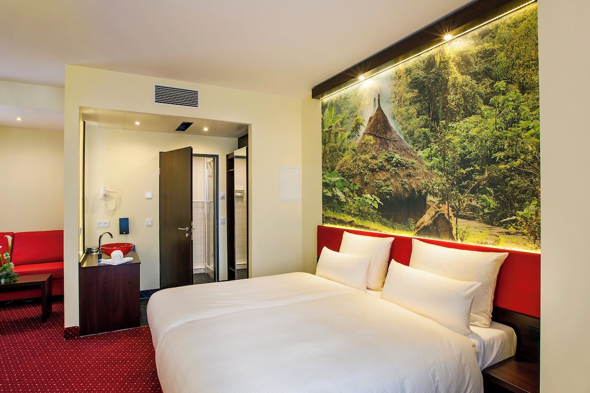 A hotel room with a large bed, a red sofa, a small table with a vase, and a painting of a forest scene.