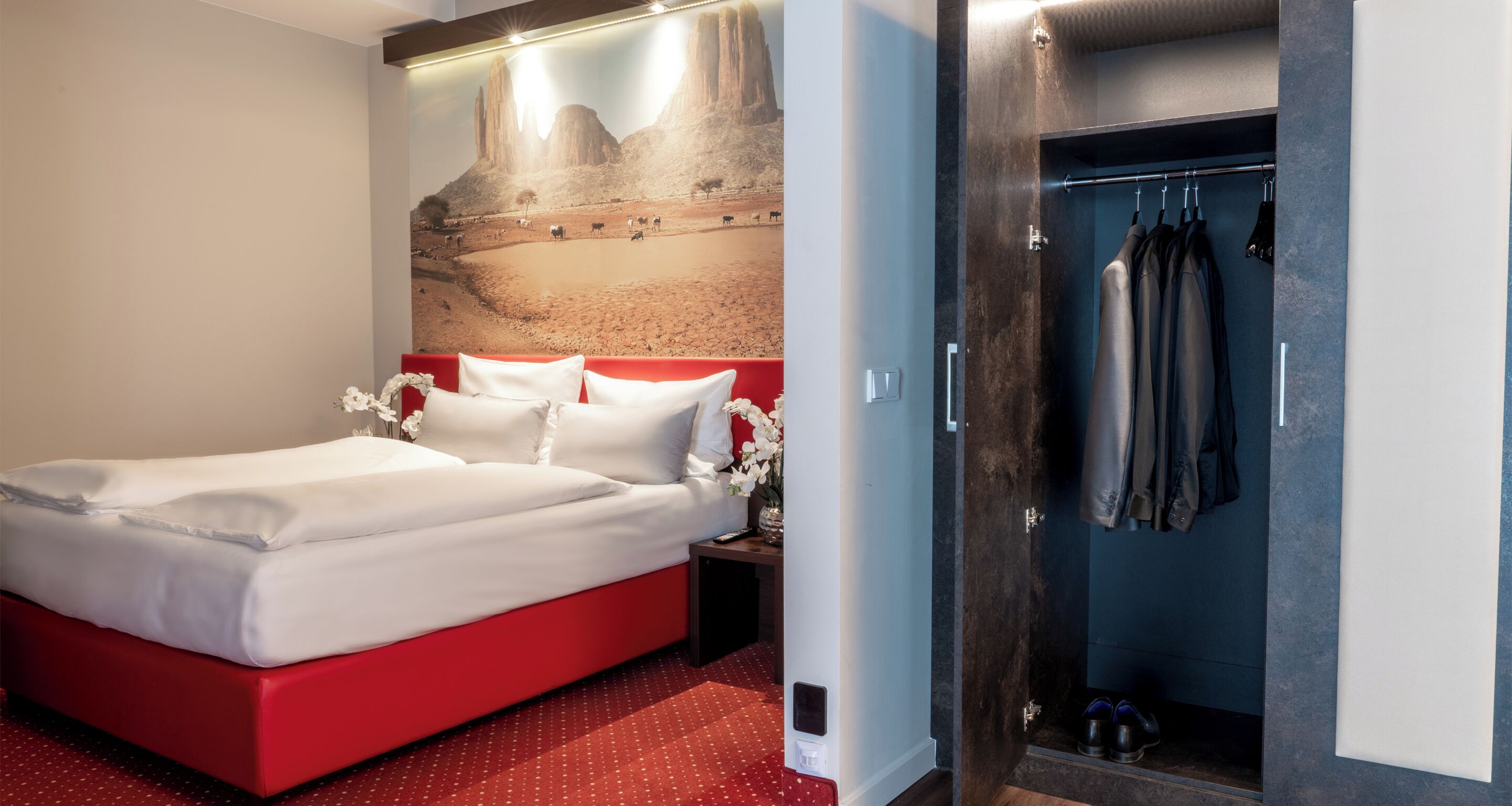 Une chambre d’hôtel avec un lit rouge, des oreillers blancs, une table de chevet ornée d’un vase de fleurs, un grand tableau au mur et un dressing où sont suspendus des vêtements et des chaussures.