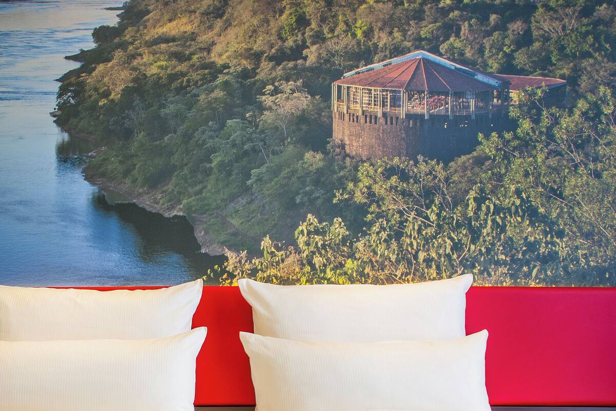 A hotel room with a red wall and two white pillows. In the background, there is a view of a river and a building on a hill.