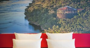 Habitación de hotel con una pared roja y dos almohadas blancas. Al fondo, se ve un río y un edificio en una colina.