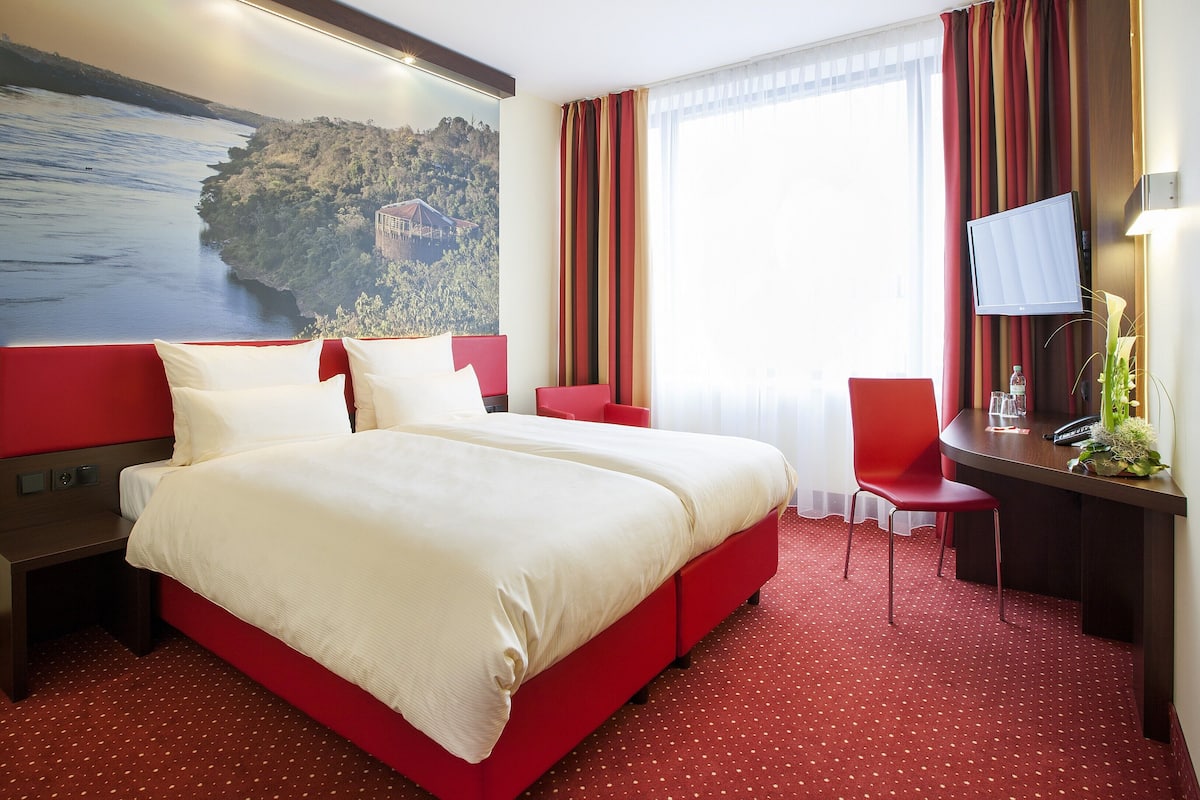 A hotel room with a large bed, a desk with a chair, a TV, and a window with curtains.