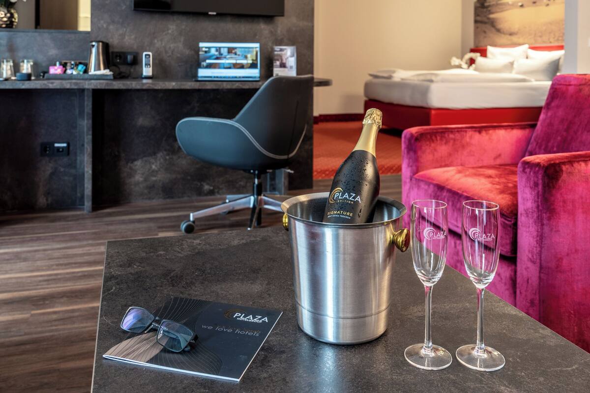 A modern hotel room with a minibar, a bed, a red velvet armchair, and a table with a bottle of champagne and two glasses.