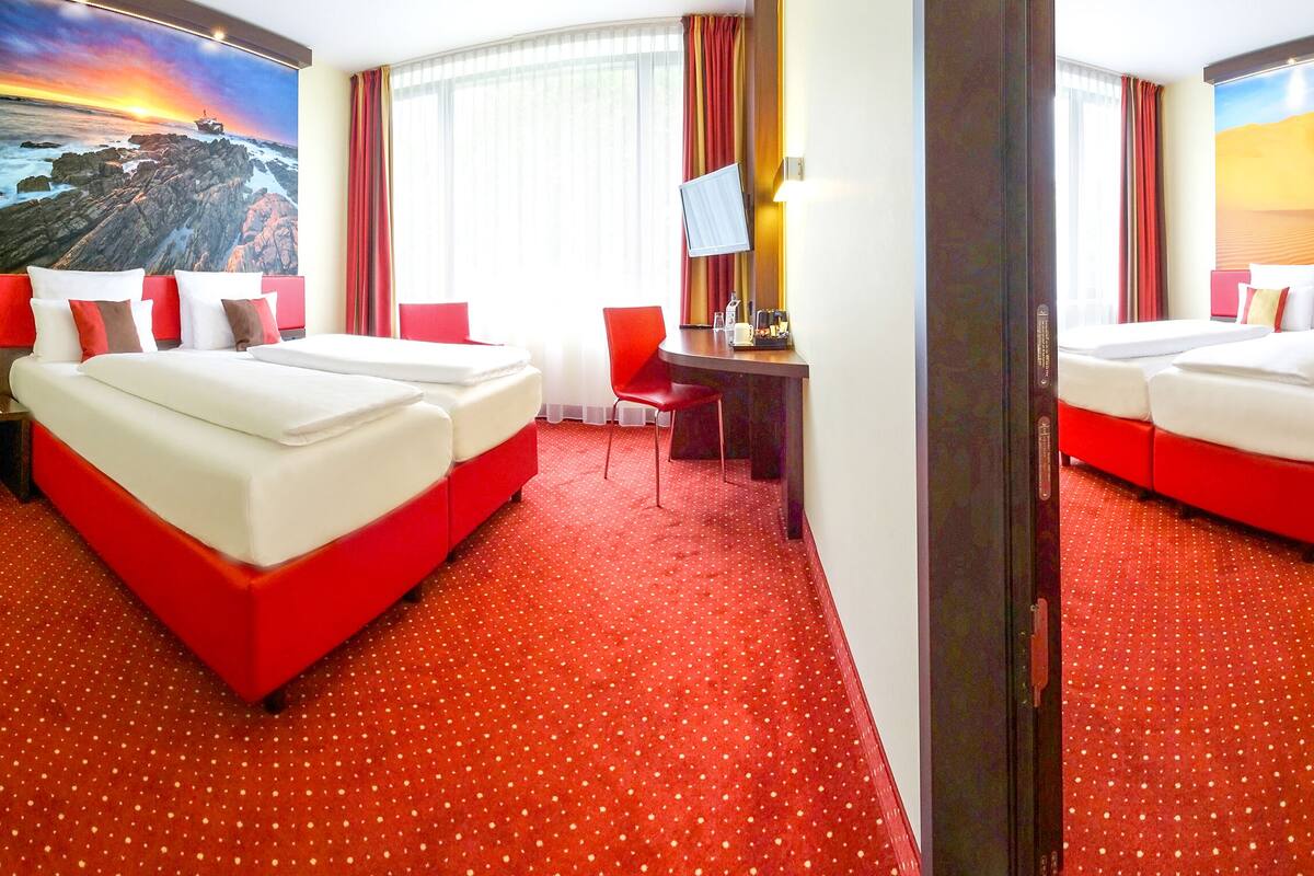 A hotel room with two beds, a desk, a TV, and a large window with curtains.