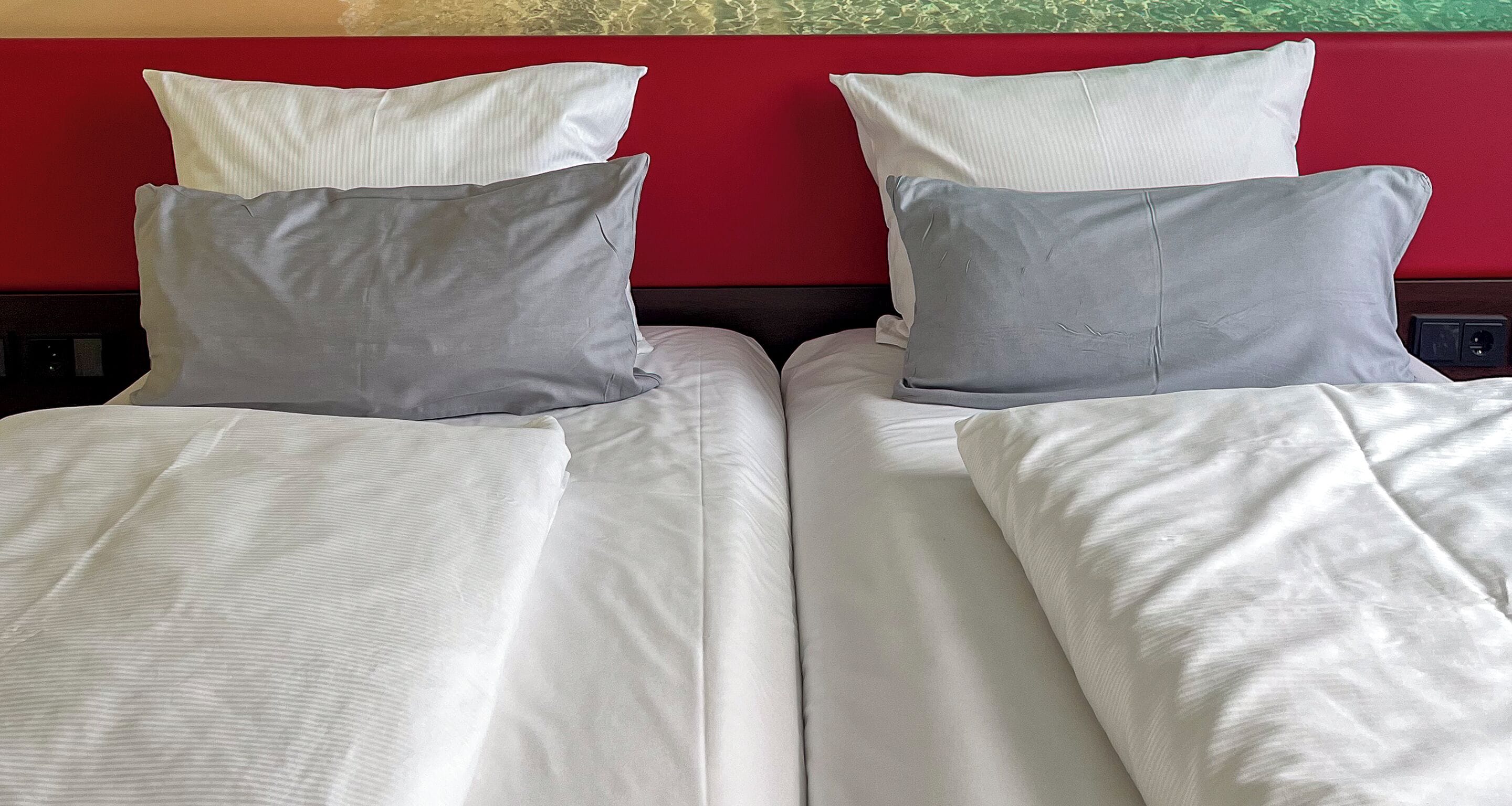 Two beds with white bedding and grey pillows, a red headboard, and a power outlet on the wall.
