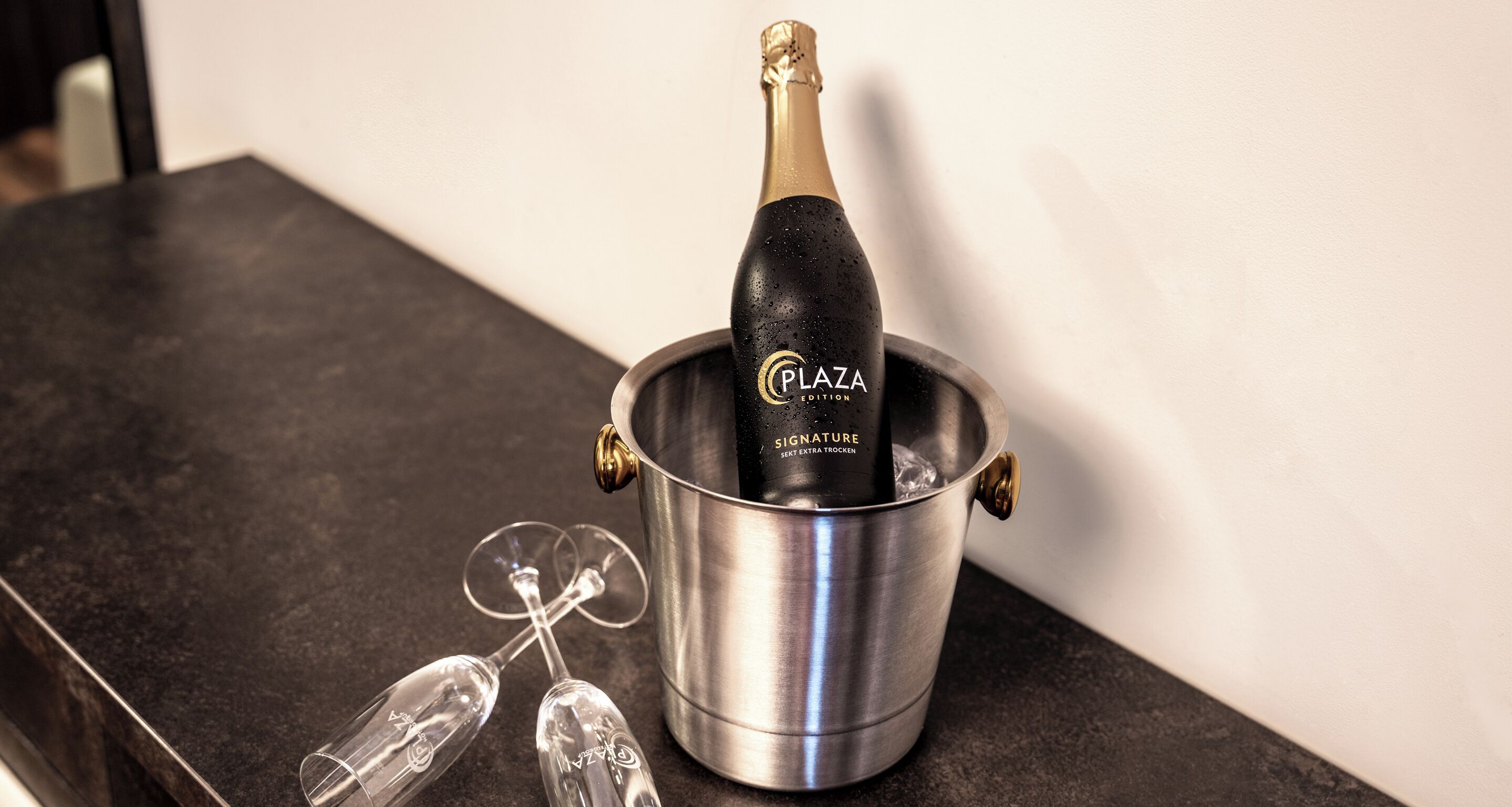 Een fles Plaza Signature Extra Trocken champagne in een metalen emmer met twee lege champagneglazen ernaast.