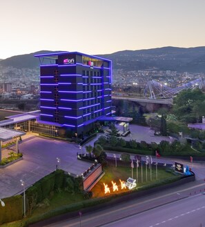 Un hotel moderno con una estructura de varios pisos, una entrada destacada y vistas a un puente y al paisaje urbano al fondo.