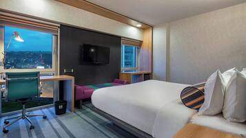 A modern hotel room with a large bed, a desk with a computer, a chair, a TV, and a window with a city view.