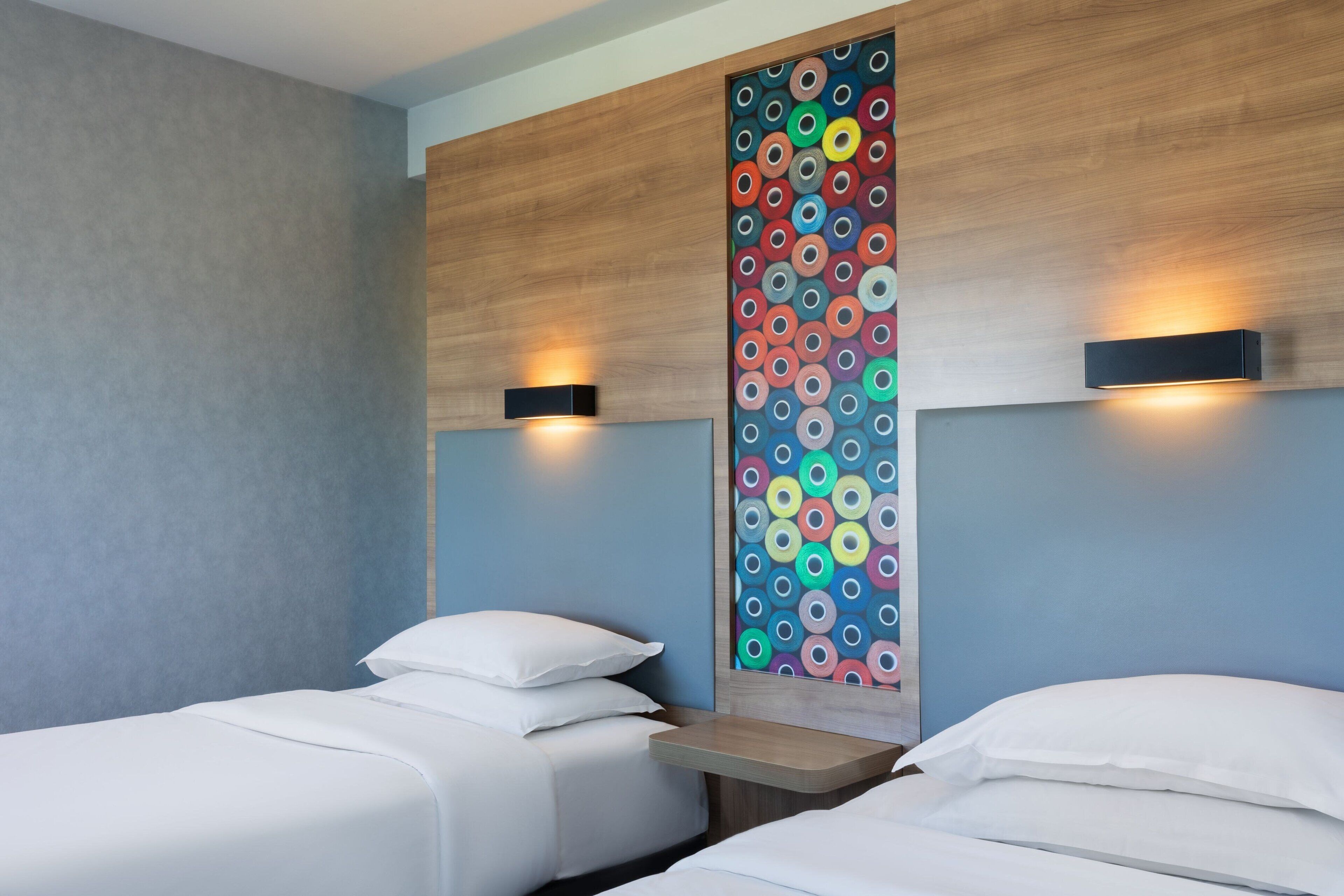 A hotel room with two beds, a wooden headboard with a colorful circular pattern, and wall-mounted lights.
