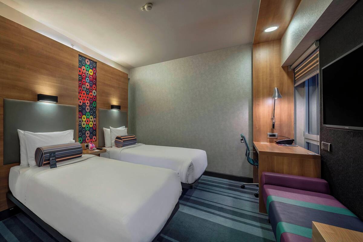 Une chambre d’hôtel avec deux lits, un bureau, une chaise, une télévision et une œuvre d’art colorée au mur.