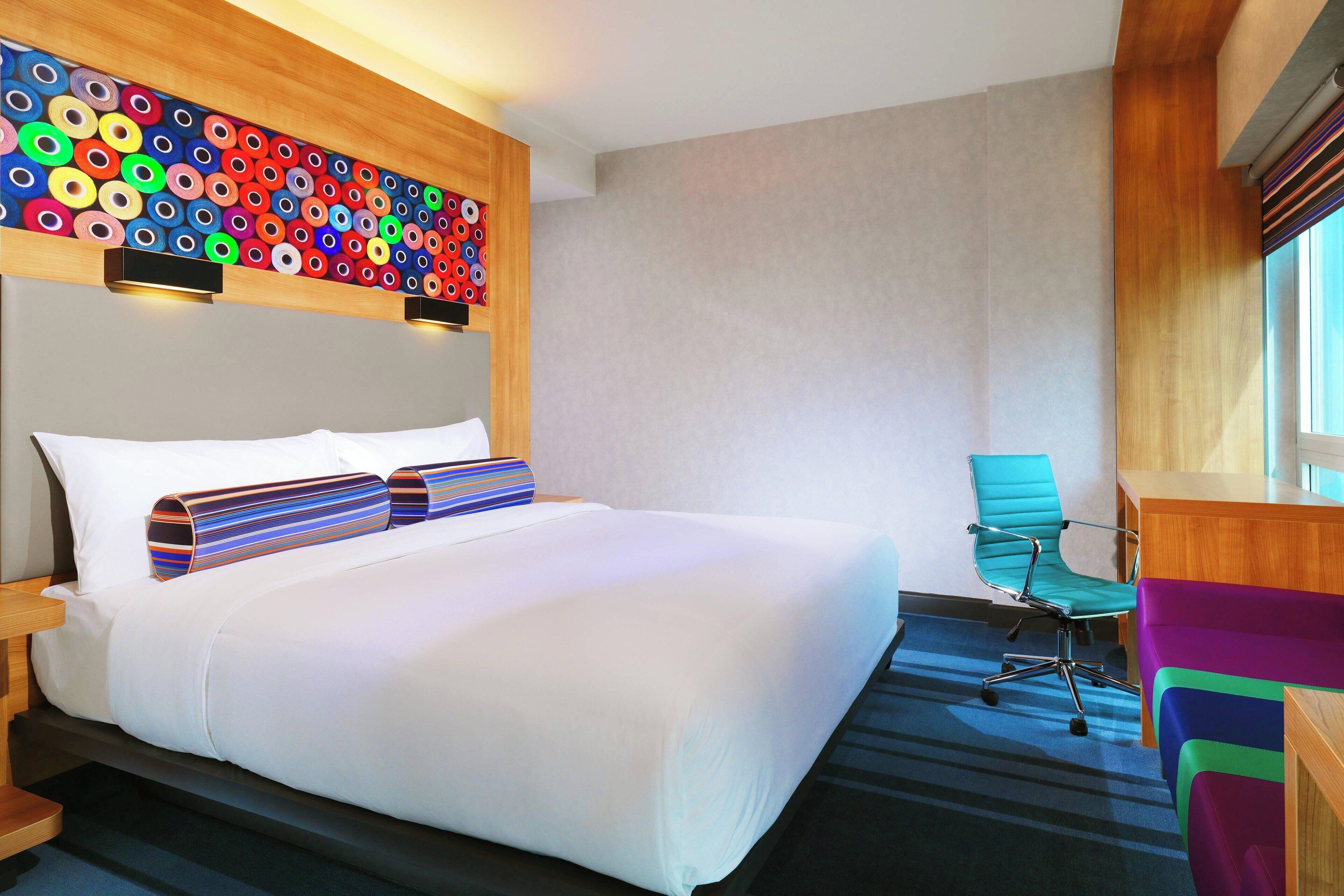 A hotel room with a large bed, a colorful wall art piece, a teal office chair, and a wooden desk.