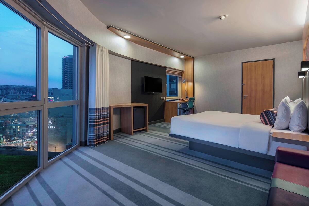Une chambre d’hôtel moderne dotée d’un grand lit, d’un bureau, d’une télévision et offrant une vue sur le paysage urbain.