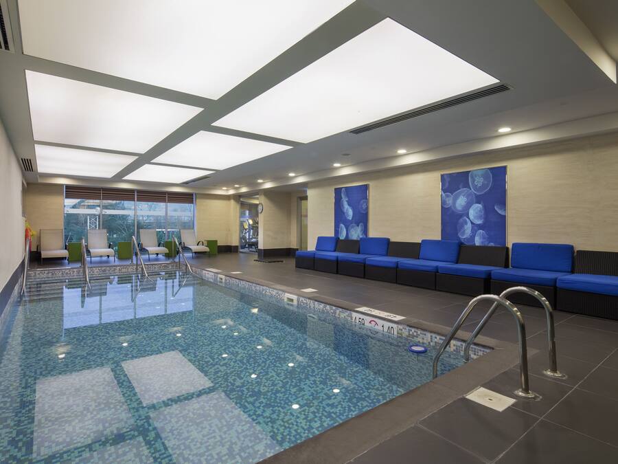 A modern indoor swimming pool with clear water, a tiled floor, and blue cushioned seating along the walls.