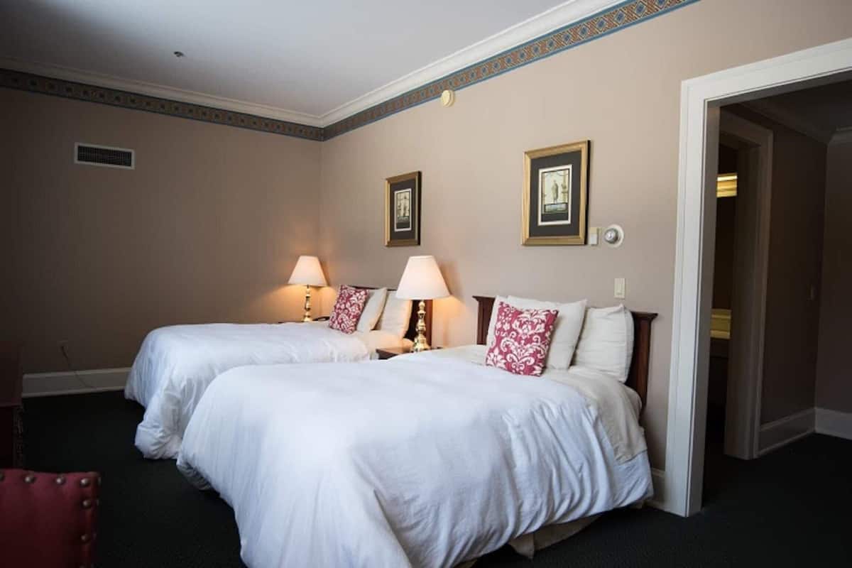 Quarto de hotel com duas camas, cada uma com roupa de cama branca e almofada estampada em vermelho e branco. Há duas luminárias de mesa, um quadro emoldurado na parede e uma porta que dá para outro quarto.