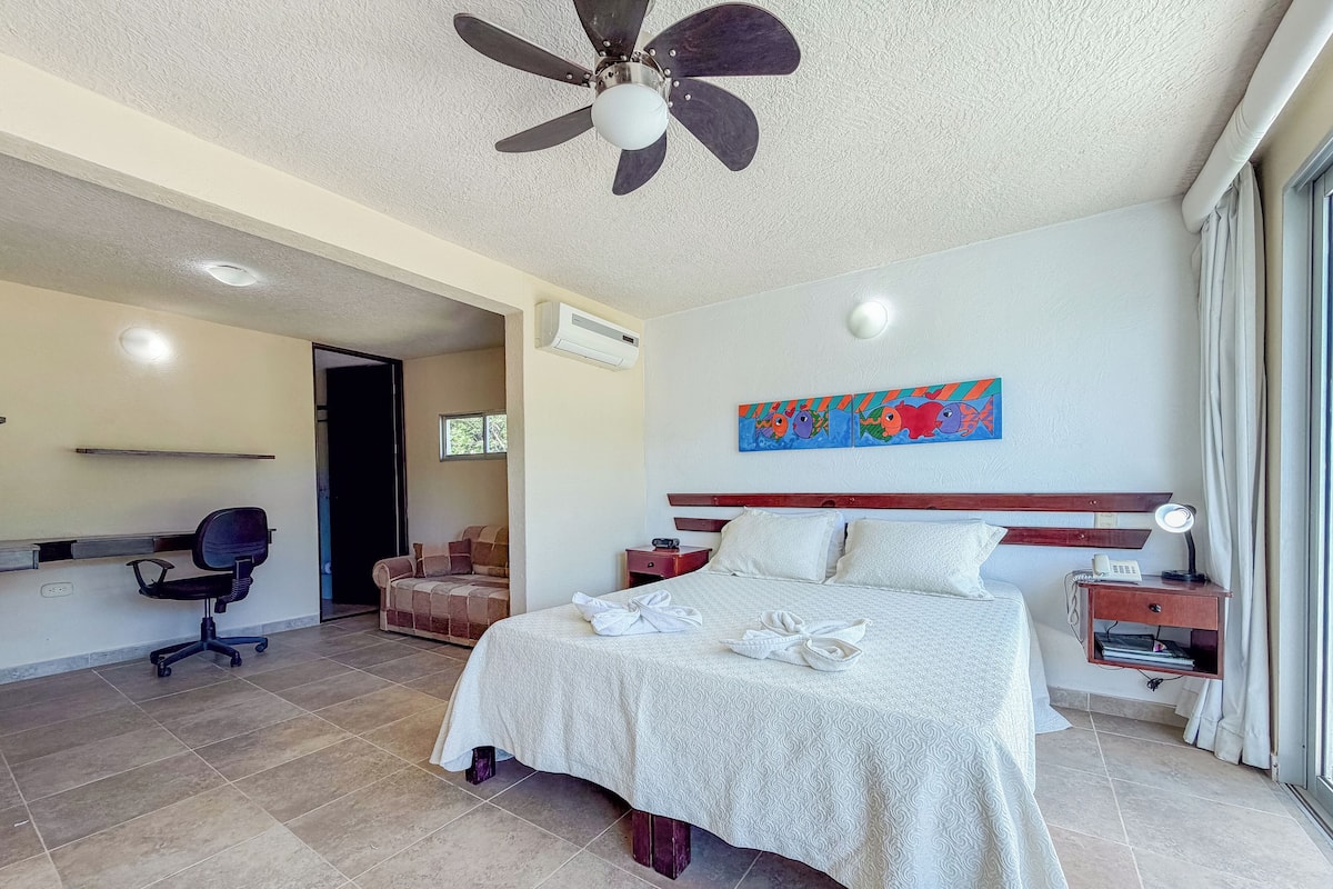 A hotel room with a bed, a desk, a chair, a ceiling fan, and a painting on the wall.
