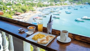 Ein Frühstücksbrett mit Rührei, Toast, Orangensaft und Kaffee auf einem Balkon mit Blick auf eine Küstenstadt, wo Boote und viel Grün zu sehen sind.