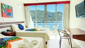 Ein Hotelzimmer mit zwei Betten, einem Schreibtisch, einem Sessel, einem Fernseher und einem großen Fenster mit Blick auf das Meer und die Hügel.
