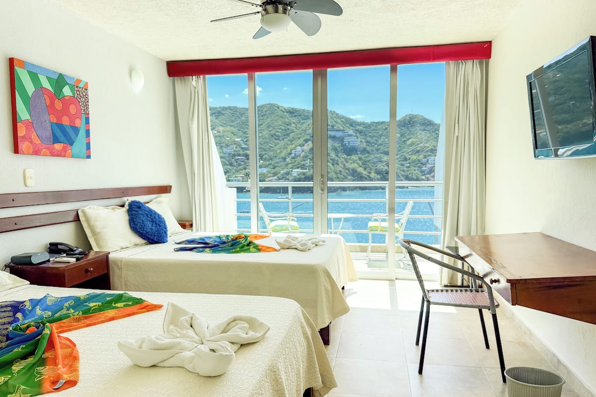 A hotel room with two beds, a desk, a chair, a TV, and a large window with a view of the sea and hills.