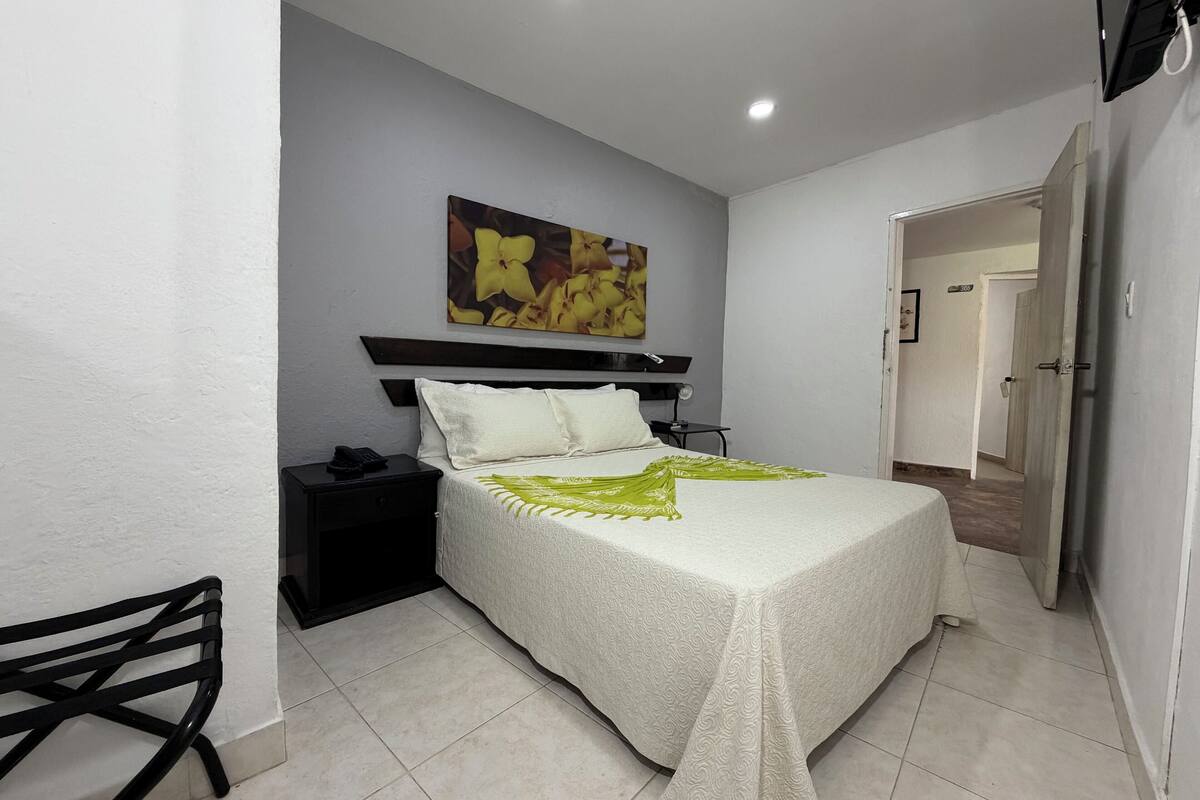 A hotel room with a bed, a nightstand, a chair, a painting on the wall, and a door leading to another room.