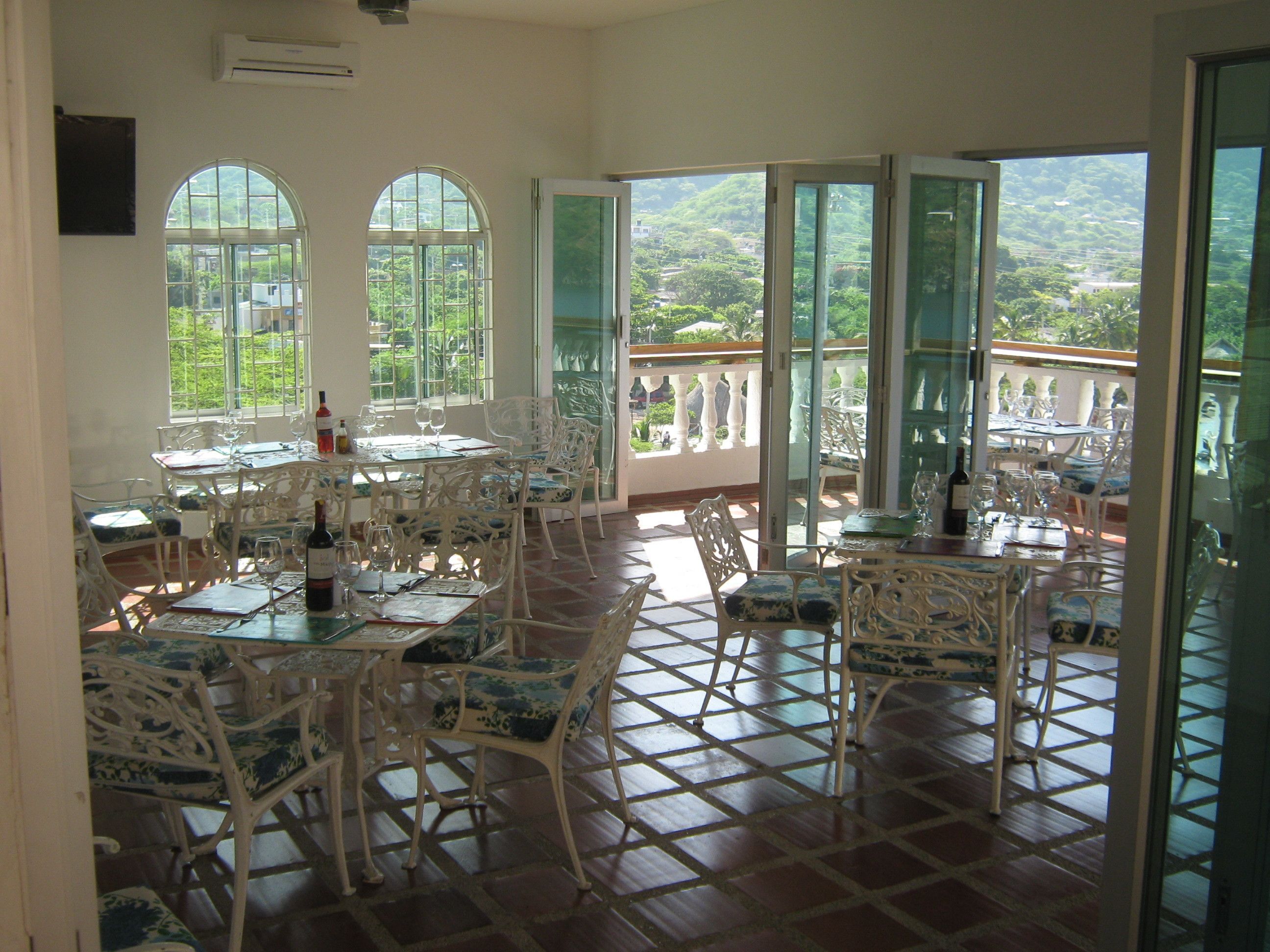 A spacious dining area with white ornate chairs and tables, large arched windows, and a view of lush greenery.