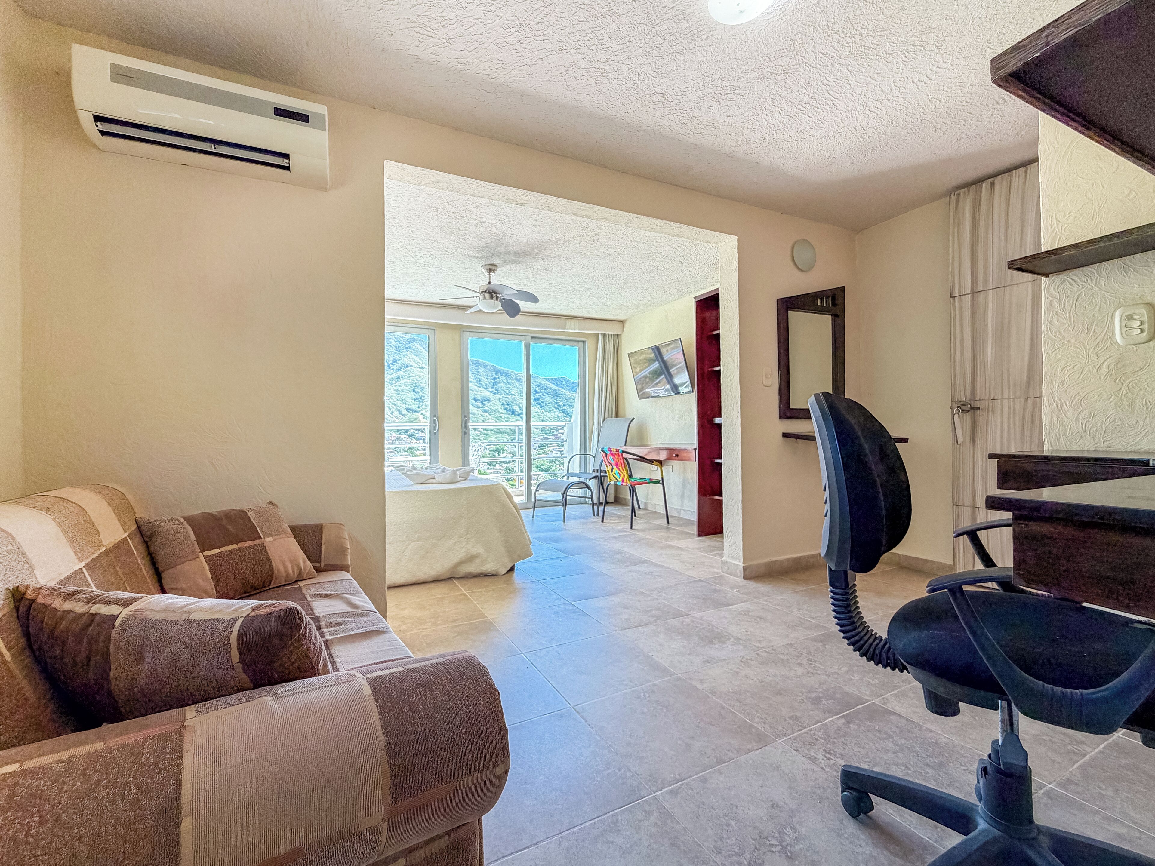 Un salon comprenant un canapé, un bureau et une chaise. On y trouve également un ventilateur de plafond, un appareil de climatisation et une vue sur un balcon avec une table et des chaises.