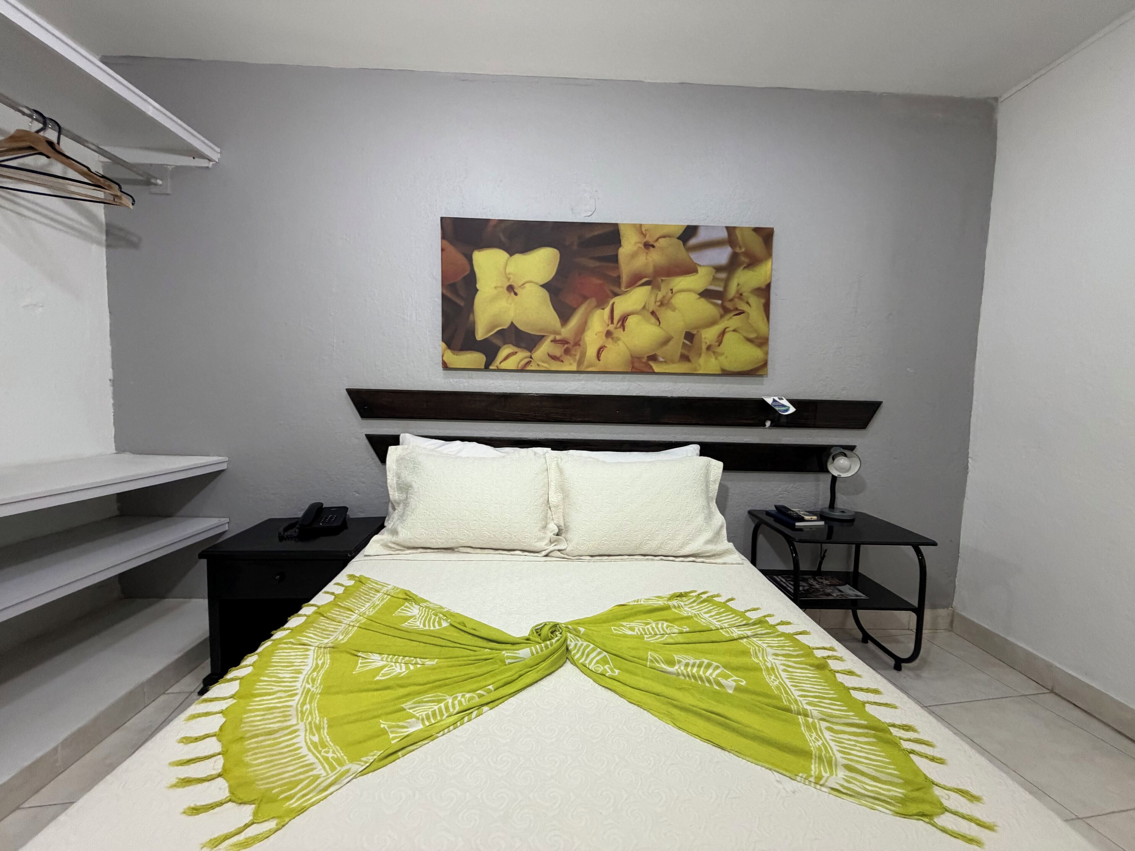 Una habitación de hotel con una cama, una mesita de noche y una lámpara de pared, además de un cuadro de flores amarillas.