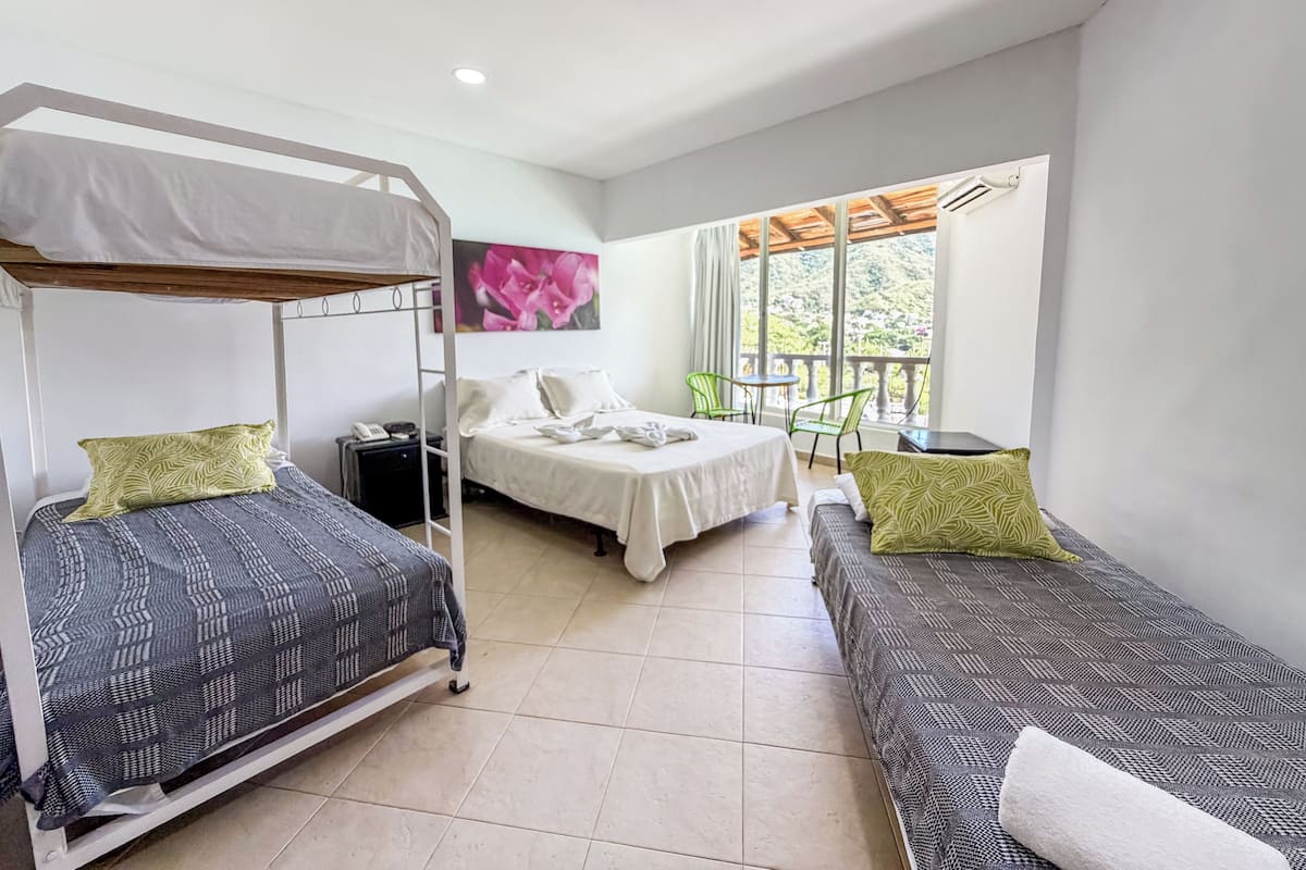 A dormitory room with bunk beds, a single bed, a small table, chairs, and a window with a view of greenery.