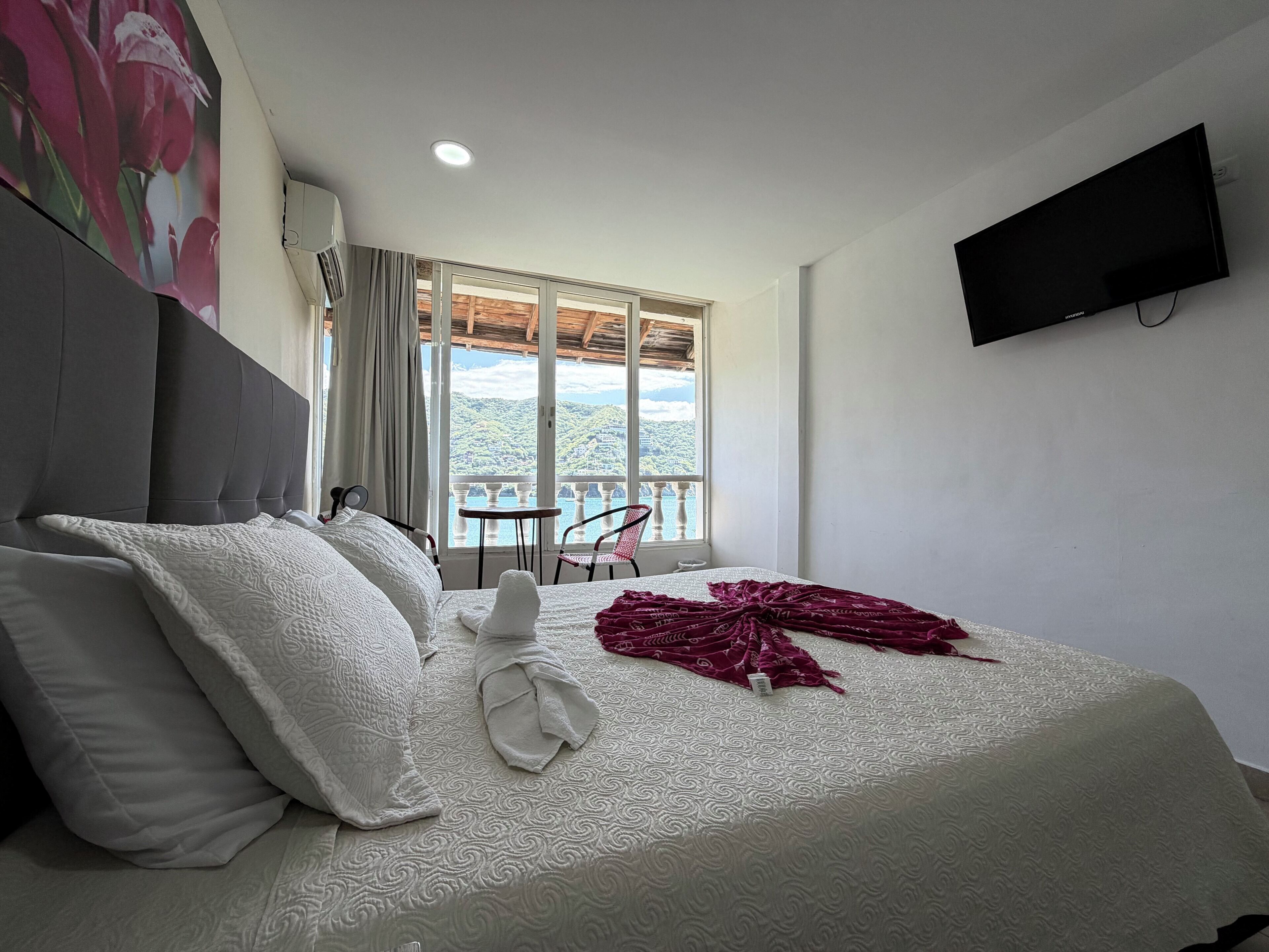 A hotel room with a large bed, a TV mounted on the wall, and a balcony with a view of the hills.