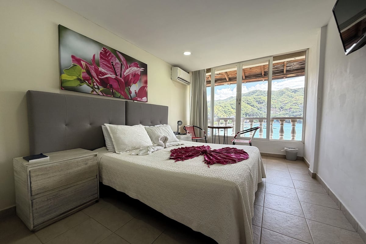 A bedroom with a bed, a nightstand, a chair, a table, a painting, and a balcony with a view.