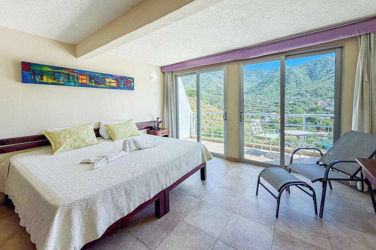 A hotel room with a bed, a chair, and a balcony with a view of the mountains.