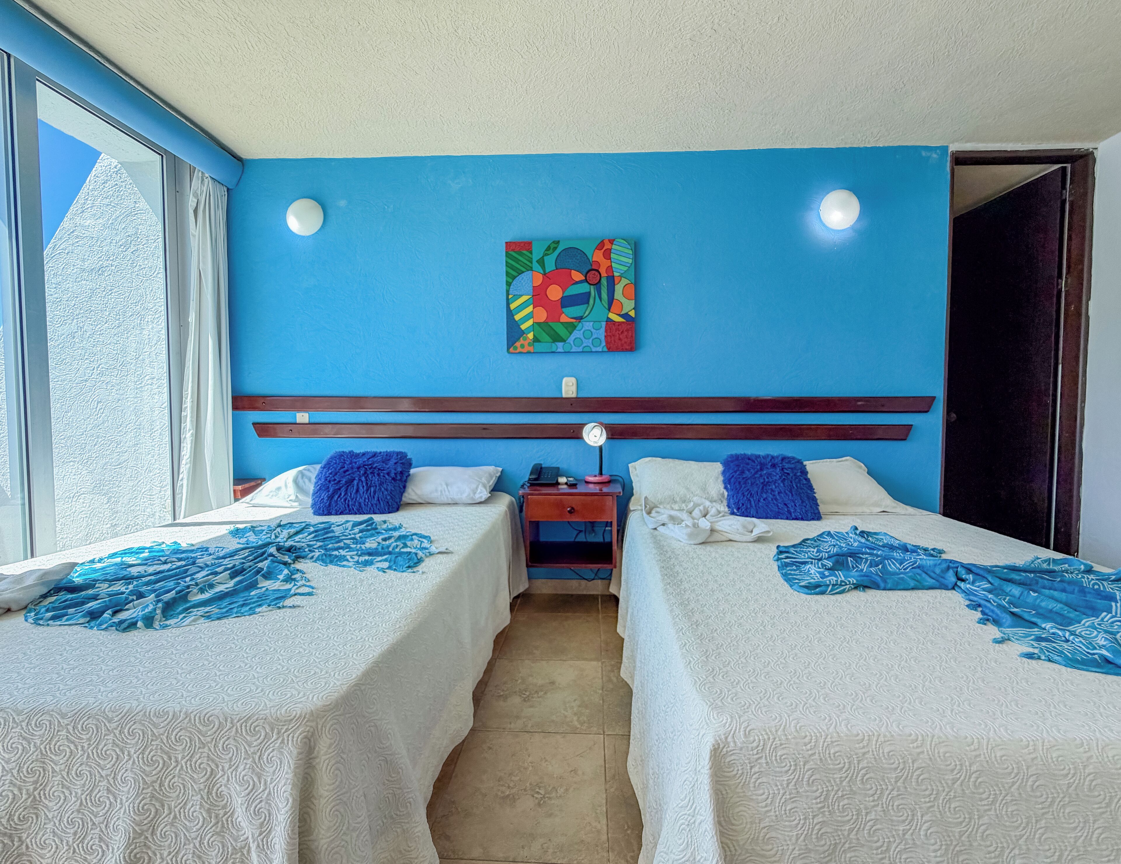 Habitación de hotel con dos camas, una pared azul, un cuadro y una ventana.
